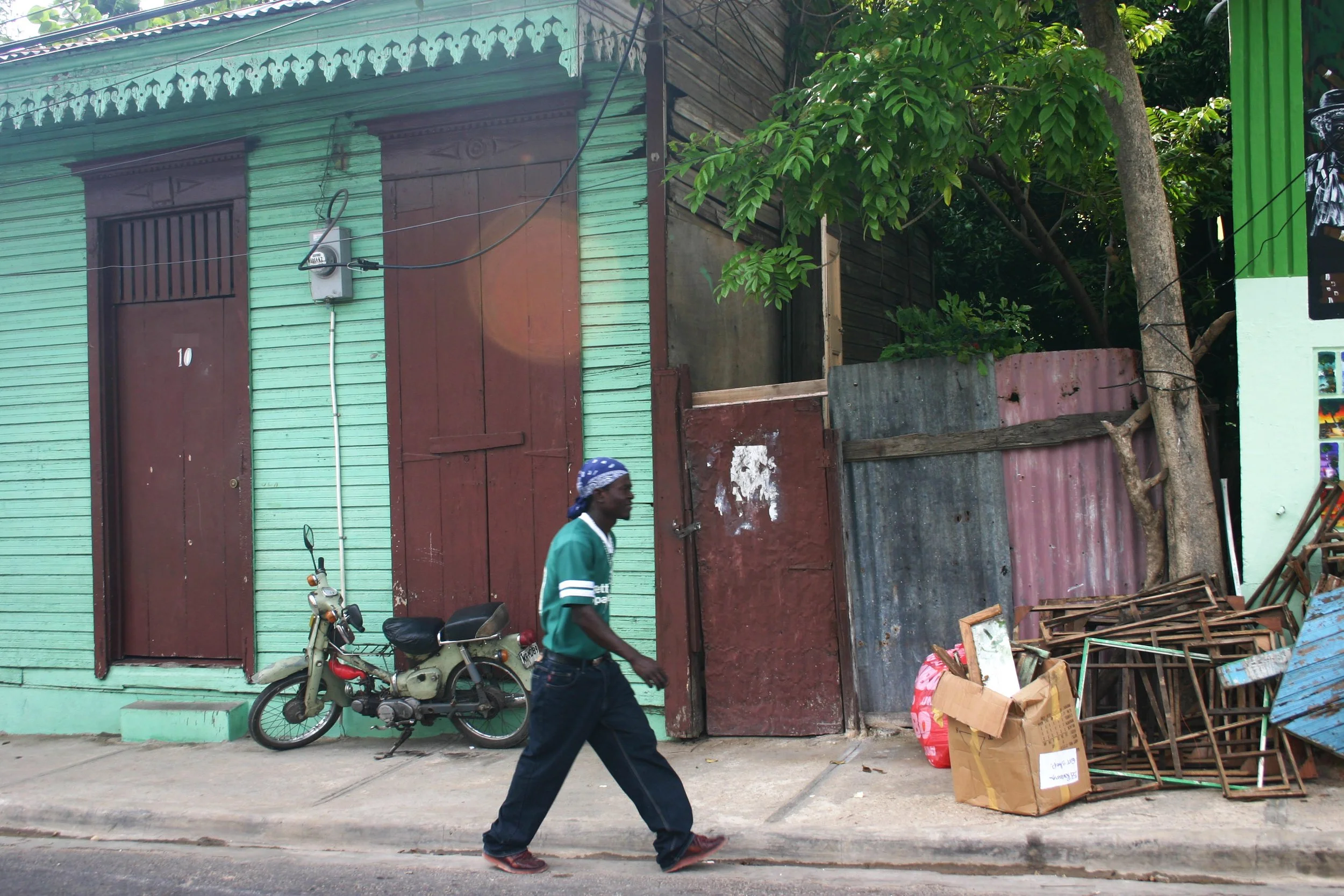 Un homme marche sur un trottoir devant une maison en bois peint en vert avec des volets en bois marron. Il y a une moto stationnée devant la maison, et un tas de métal recyclé ou abandonné et des cartons empilés à côté de la maison. Un arbre avec des