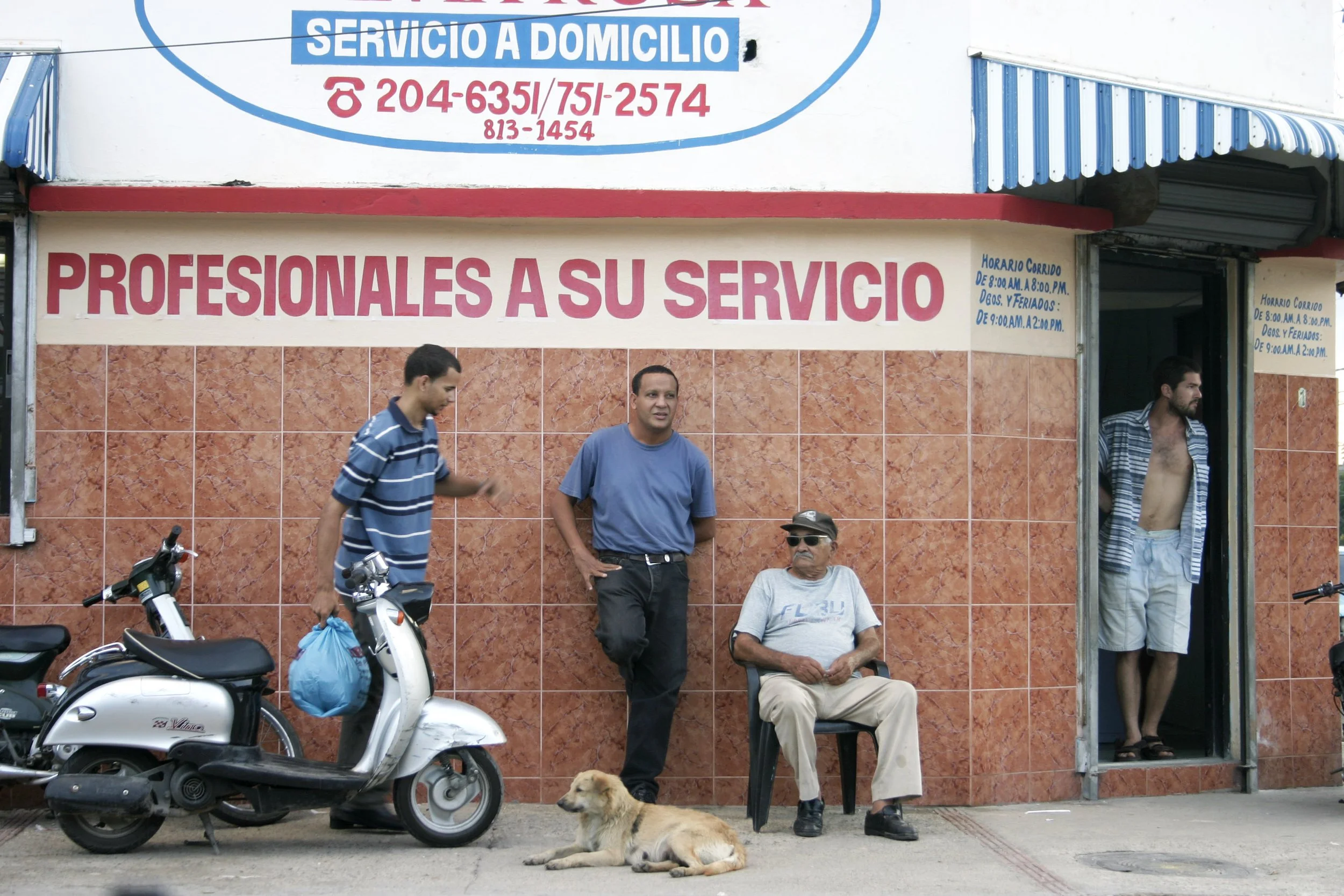 Un groupe de personnes attendant devant une boutique avec un chien couché sur le trottoir, une moto garée à côté, et un homme assis portant des lunettes de soleil, le tout ensoleillé avec un fond de mur en carrelage orange et un panneau en espagnol.