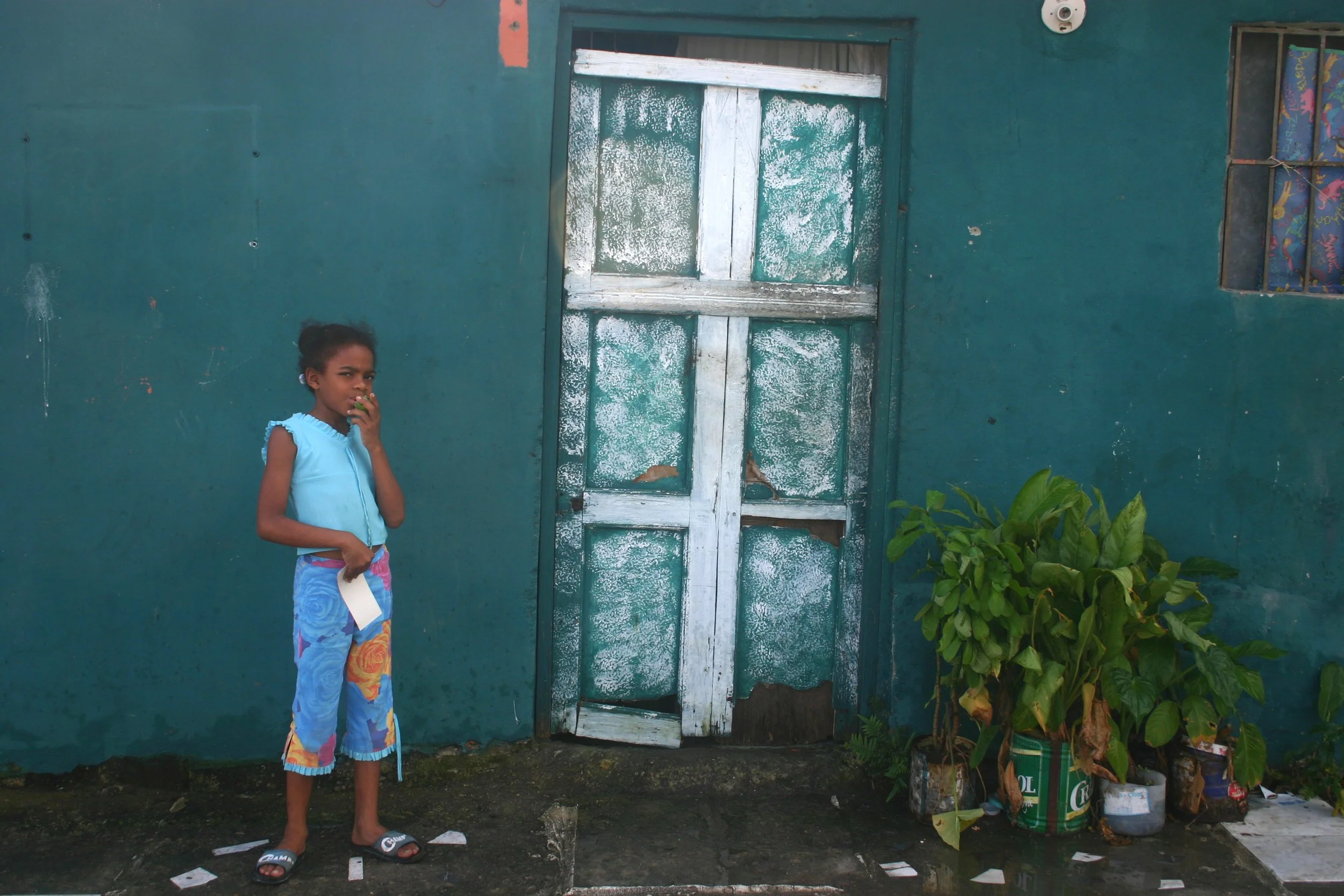 Une jeune fille en vêtements colorés se tient devant une maison aux murs bleus et une porte en bois blanche délabrée. Elle porte des sandales et tient un morceau de papier, semblant manger un fruit ou un bonbon. À droite, il y a des pots de fleurs av