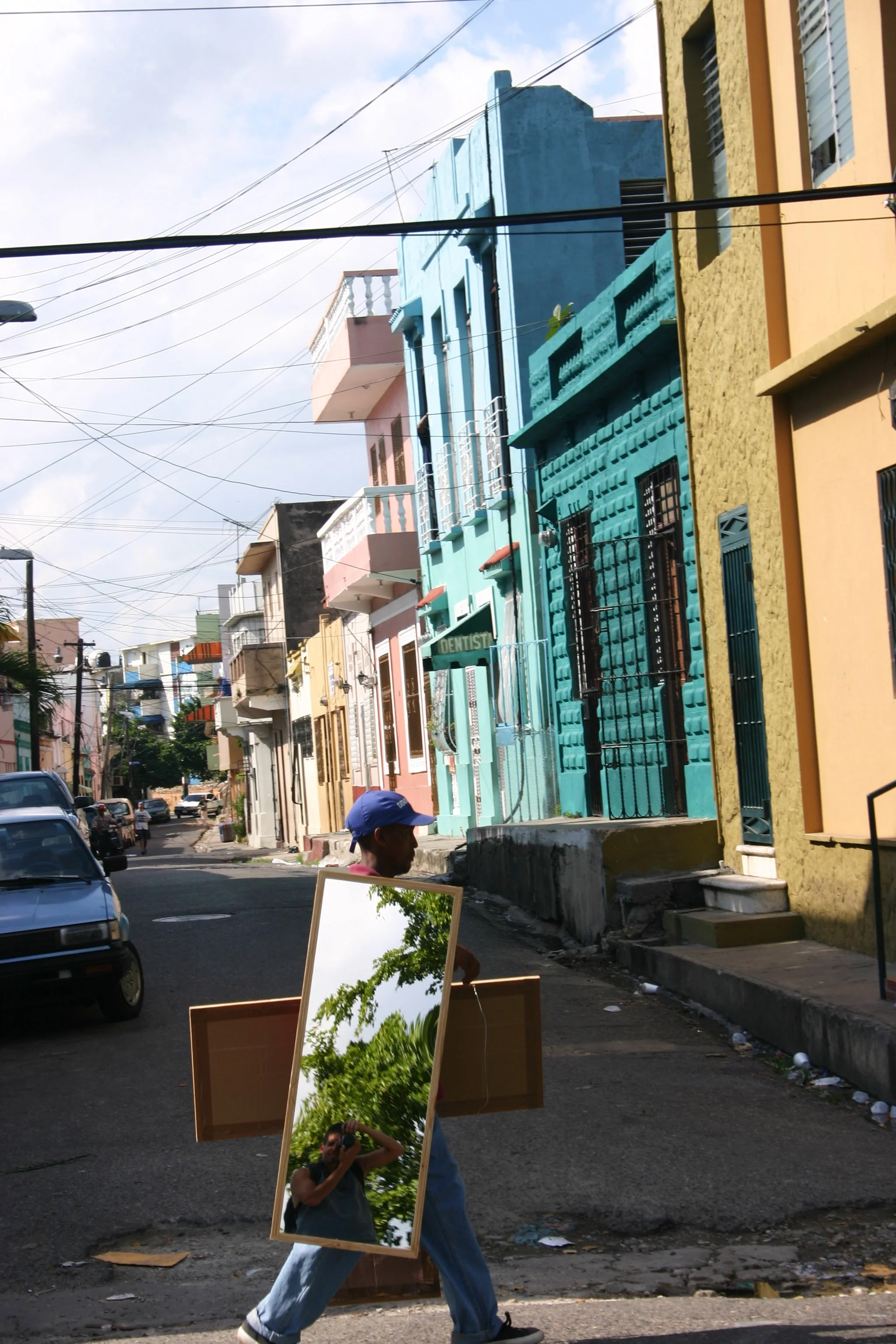 Un homme marche dans une rue colorée avec un grand miroir porté dans le dos, reflétant des arbres et le ciel, entouré de bâtiments colorés et de voitures stationnées.