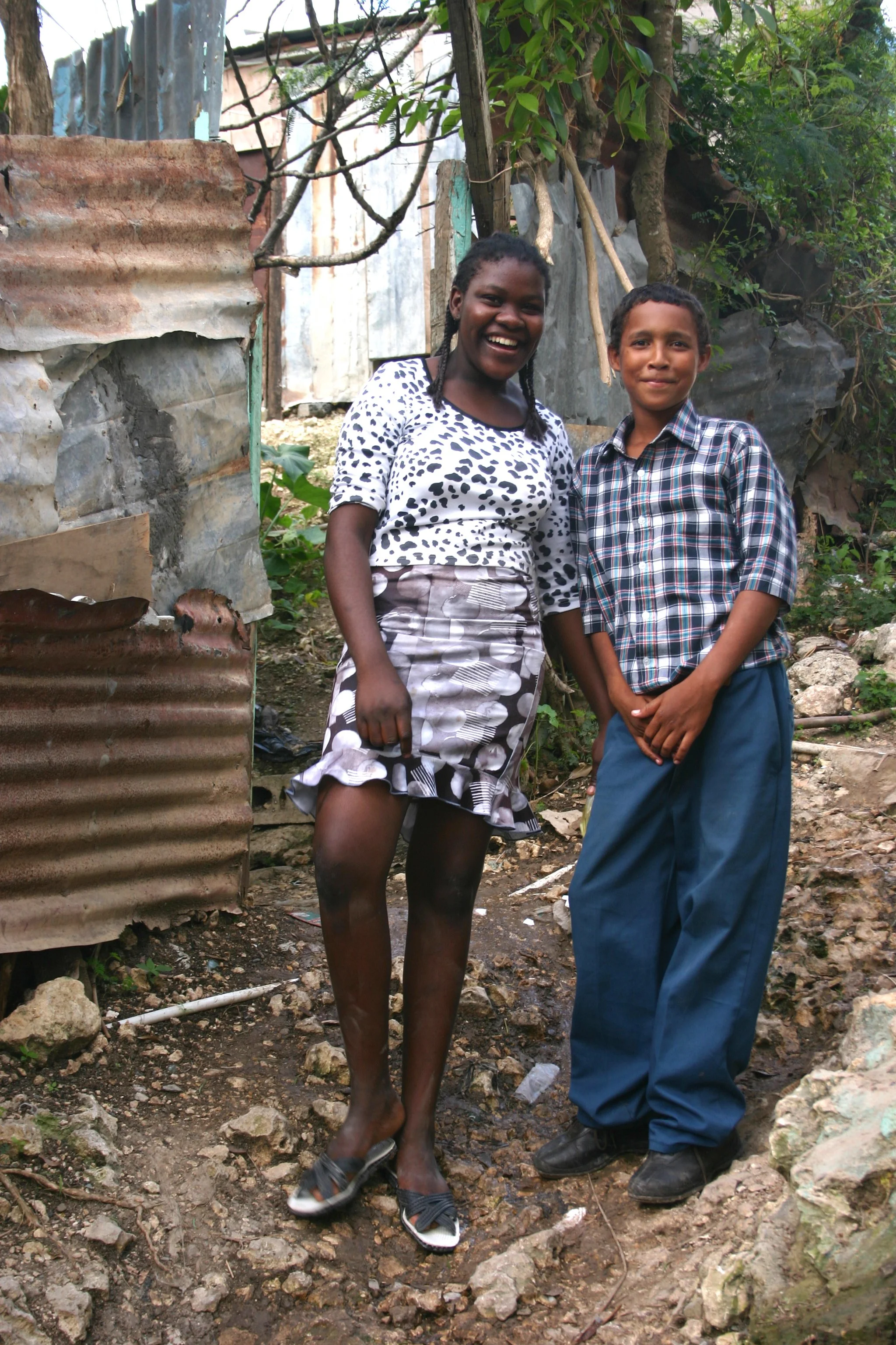 Deux enfants souriants debout côte à côte dans une zone urbaine défraîchie. La fille porte une robe à motifs et des sandales, tandis que le garçon porte une chemise à carreaux et un pantalon ample. En arrière-plan, il y a un mur en tôle ondulée et de