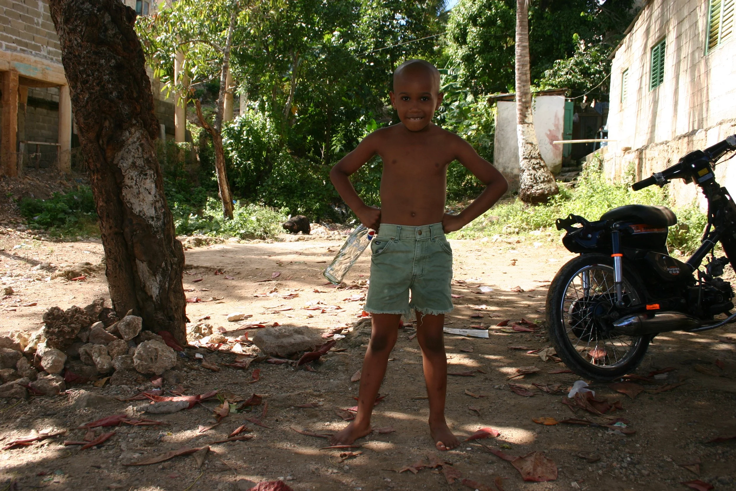 Un jeune garçon torse nu, portant un short en jean, se tient avec les mains sur les hanches dans une zone extérieure ombragée, avec un moteur stationnaire à côté, entouré d'arbres et de bâtiments en arrière-plan.