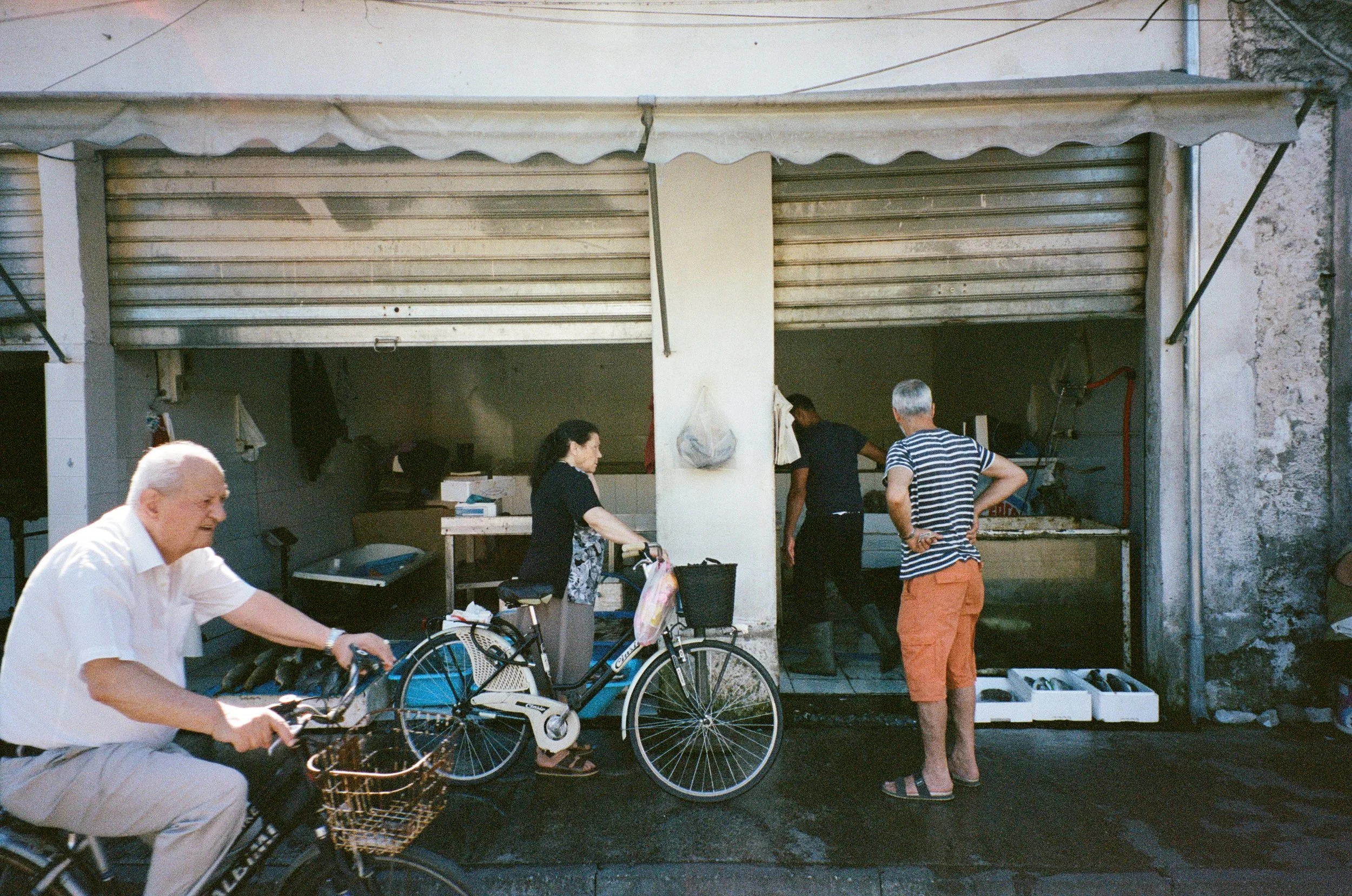 Une petite boutique avec des rideaux en métal partiellement ouverts. Plusieurs personnes attendent ou achètent, y compris un homme âgé à vélo, une femme avec un vélo électrique, et deux hommes, un en maillot rayé et un autre en noir, à l'entrée de la