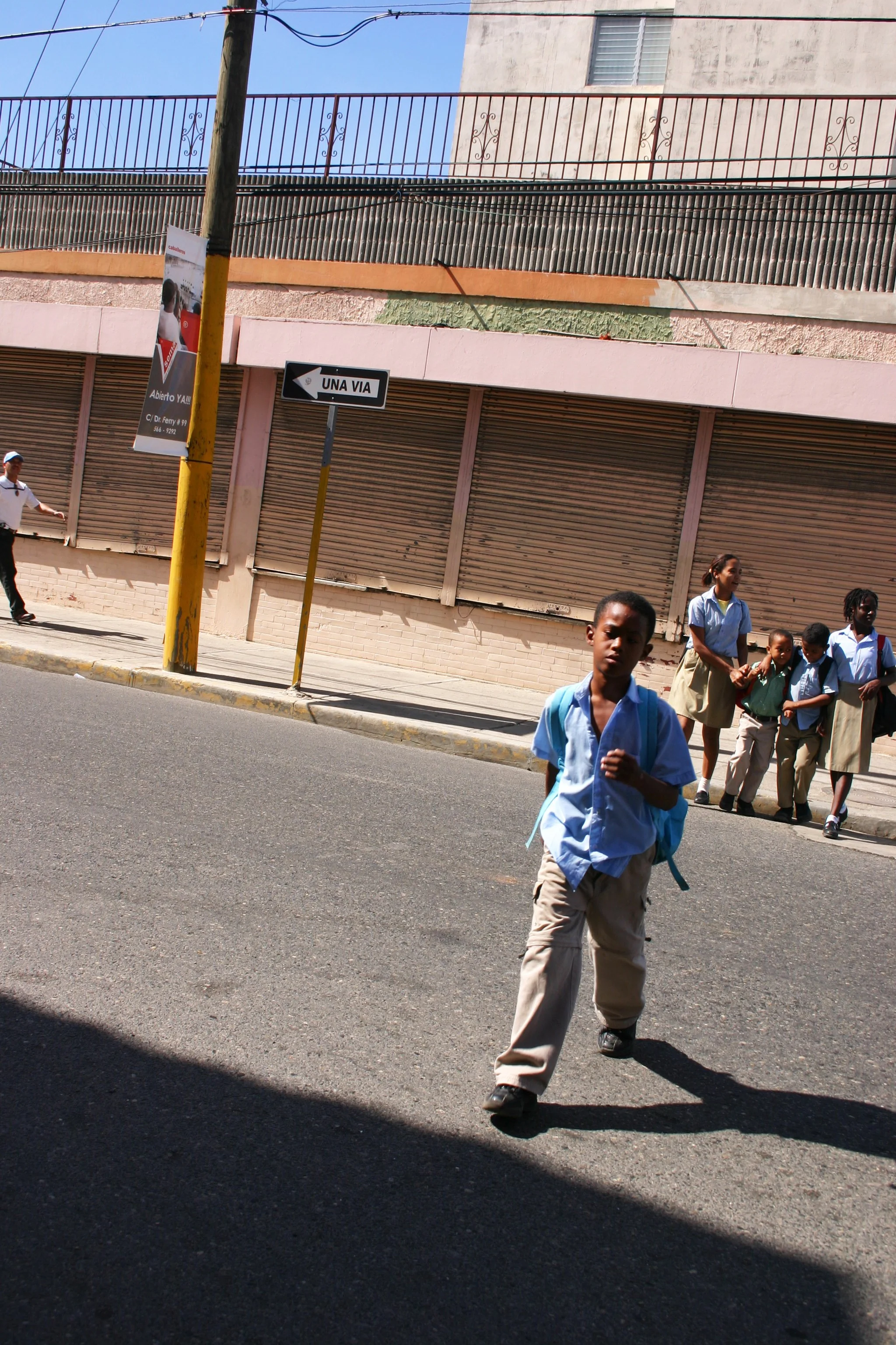 Un groupe d'enfants en uniforme scolaire traversant la rue dans une ville ensoleillée, avec un panneau de direction indiquant 'UNA VÍA'.