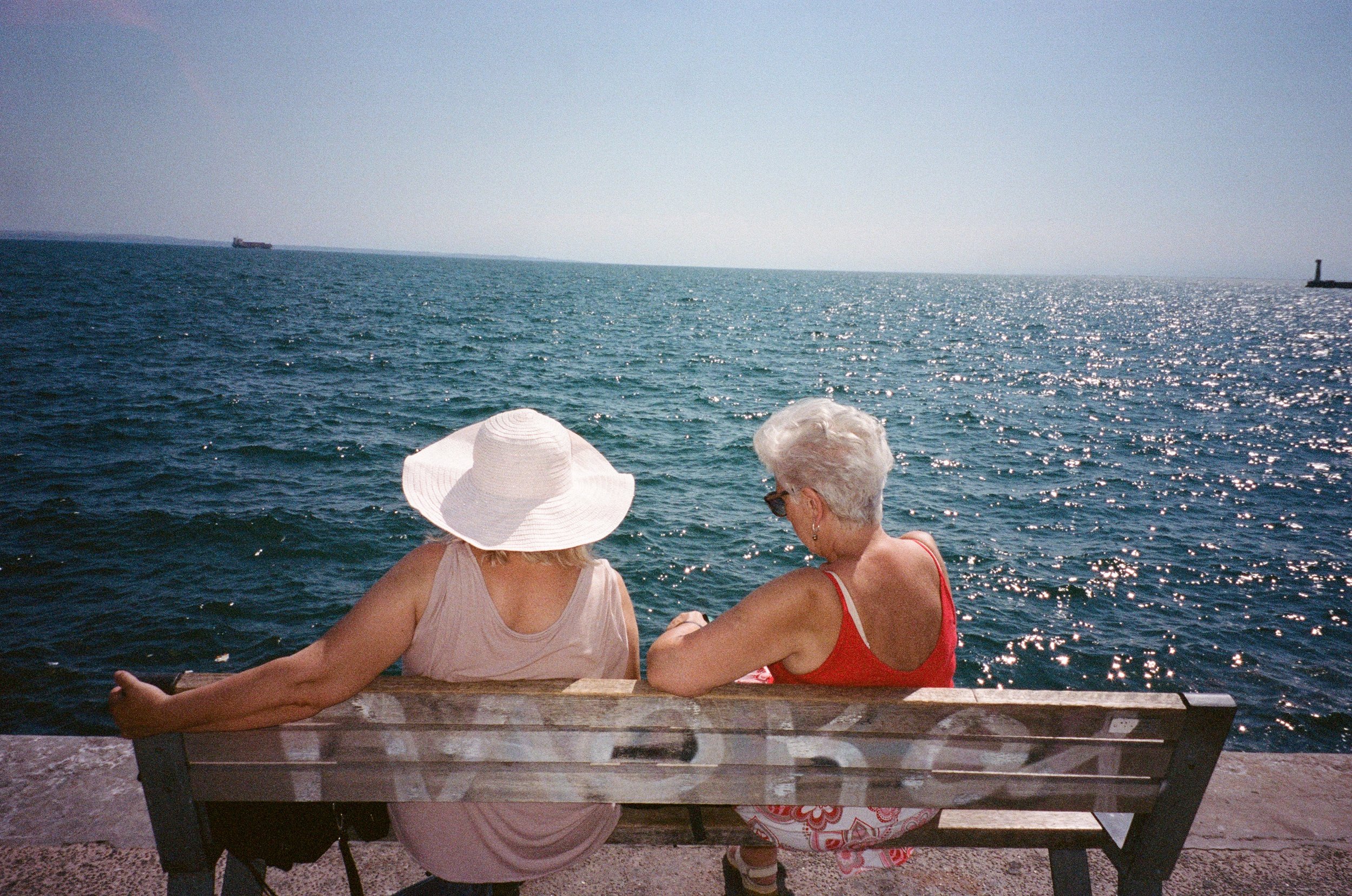 Deux femmes assises sur un banc en regardant la mer, l'une porte un chapeau blanc, elles semblent discuter ou profiter du paysage.