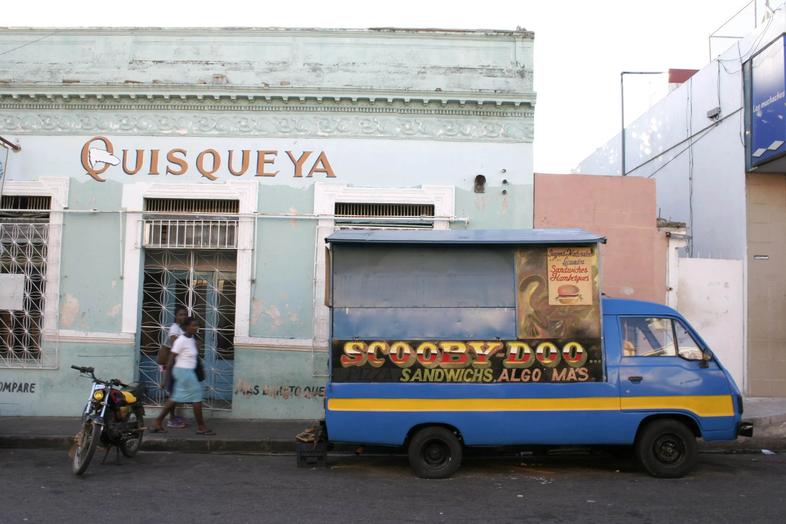 Un camion de nourriture de rue coloré bleu avec une enseigne indiquant Scooby Doo, Sandwichs et Algo Más garé devant un bâtiment avec le nom 'QUISQUEYA' peint en rouge. Deux personnes marchent sur le trottoir et une moto est stationnée devant le cami