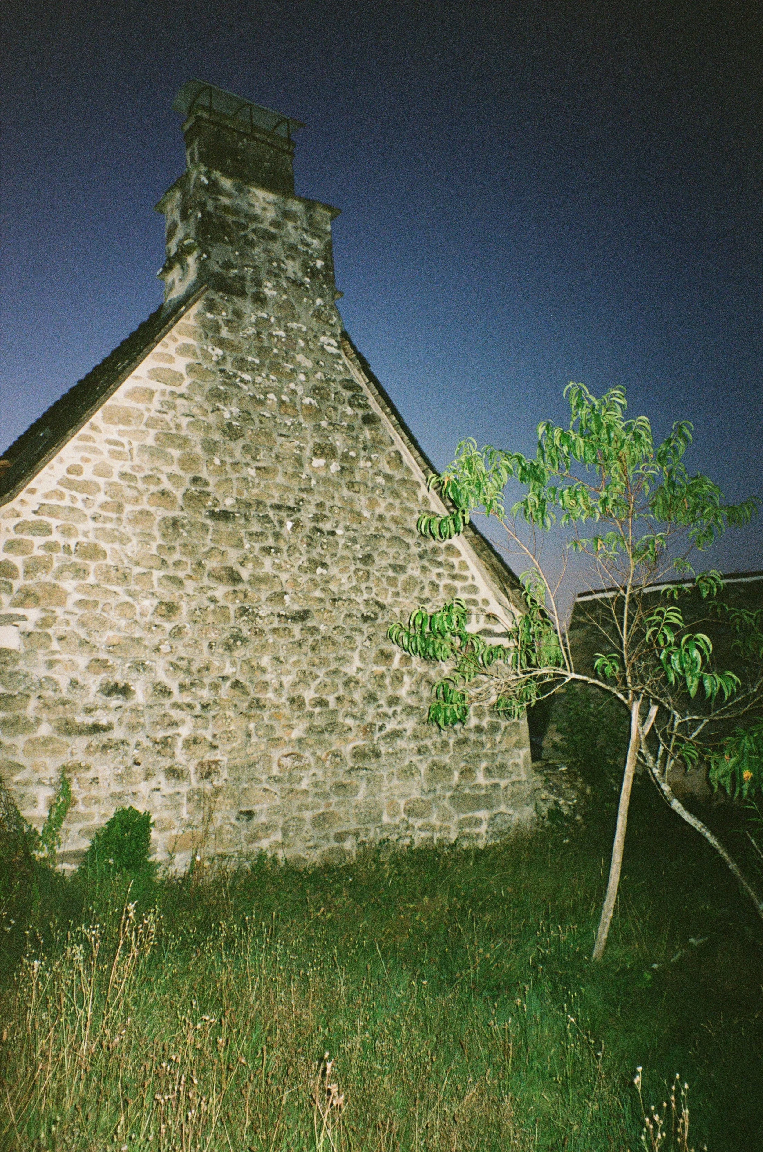 Une maison en pierre avec une cheminée, prise de nuit, avec un arbre et de l'herbe au premier plan.