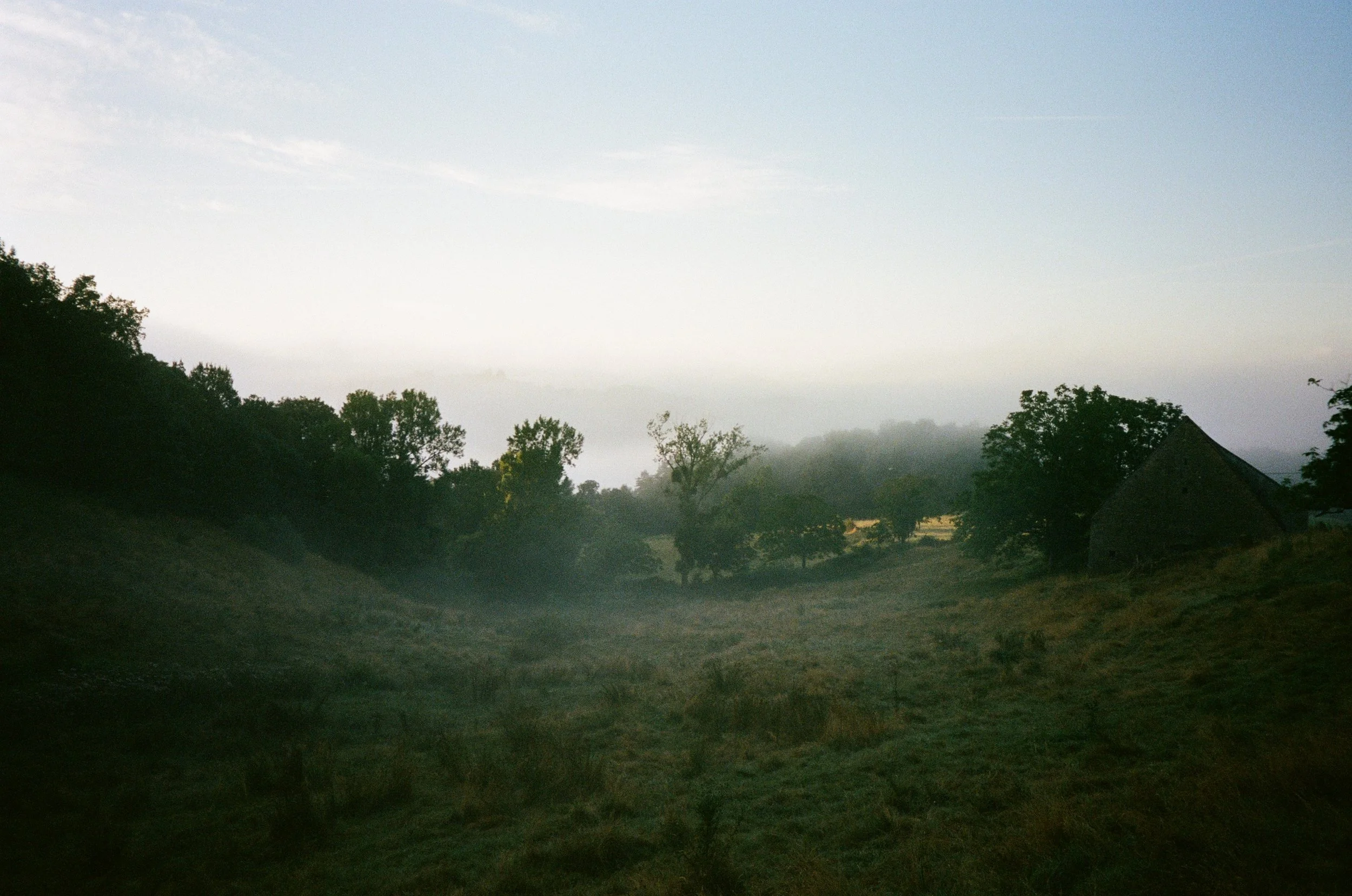 Paysage rural avec des collines et des arbres, au lever du soleil ou au crépuscule, avec une maison en pierre sur la droite.