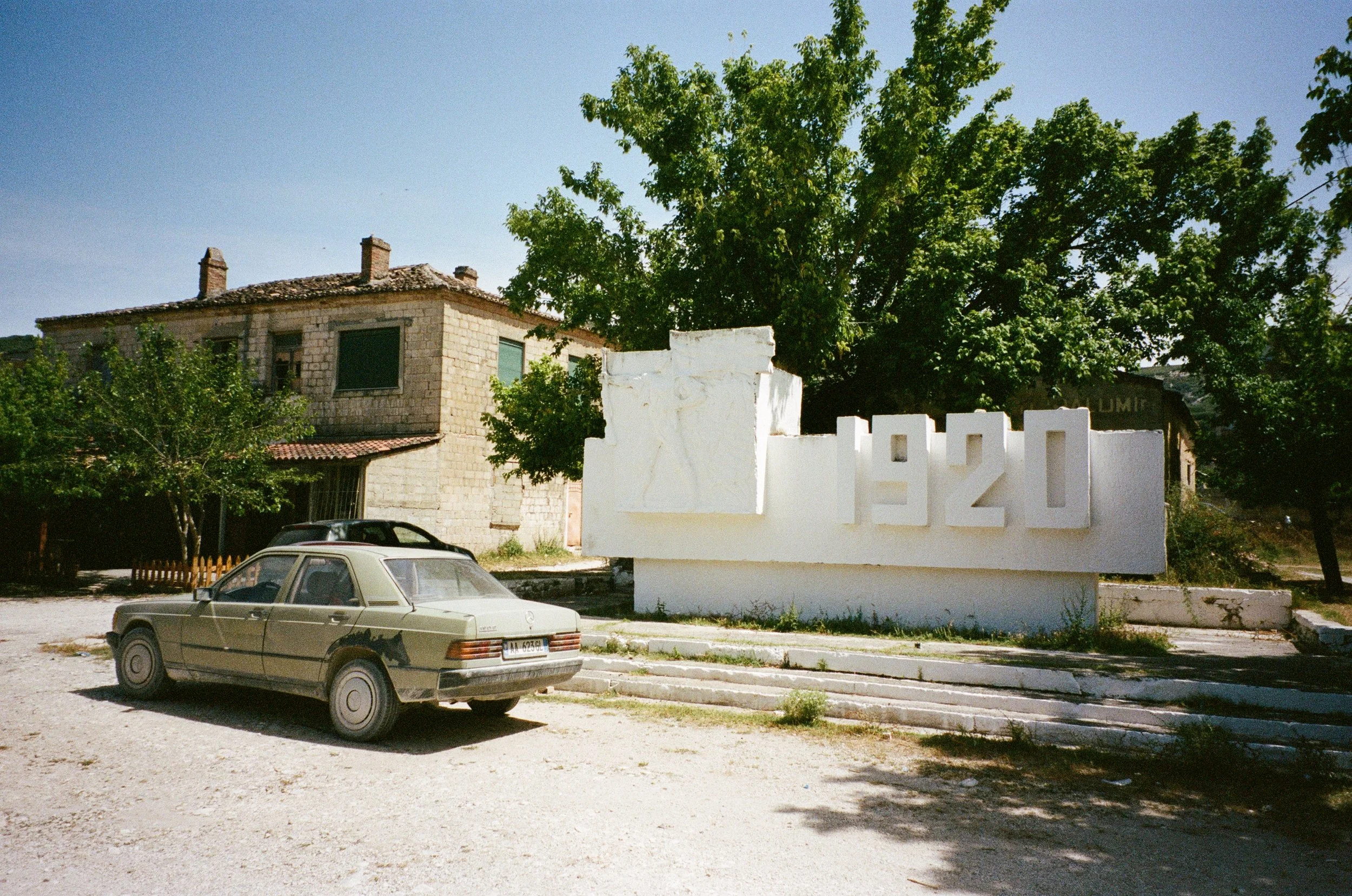 Un vieux bâtiment en pierre avec un toit en tuiles rouges, une voiture ancienne beige devant un grand panneau blanc avec la date 1920 et une sculpture en relief d'une croix sur le panneau. Arbres verts et un ciel clair en arrière-plan.