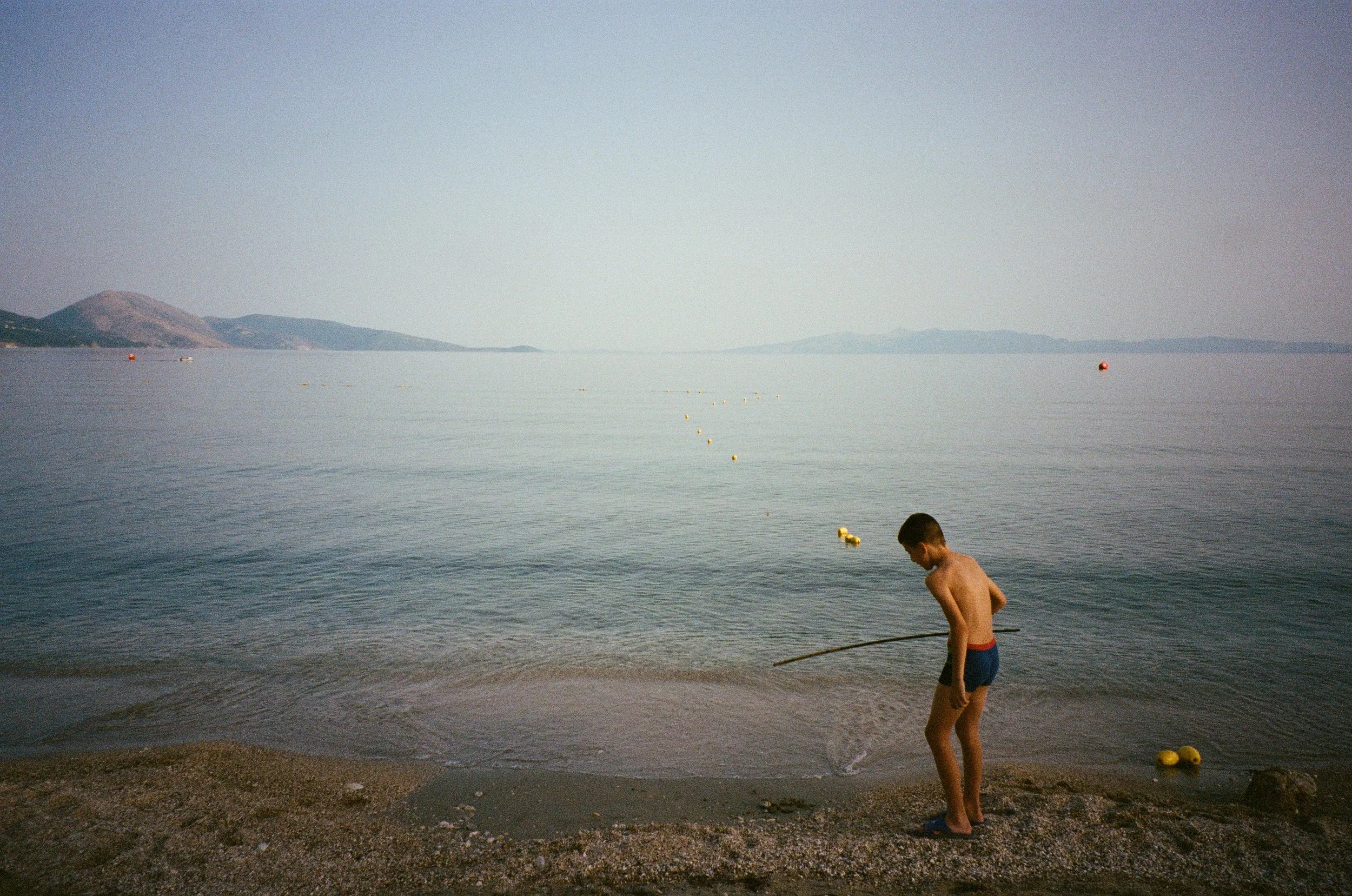 Un enfant en maillot de bain court au bord de l'eau avec une canne à pêche, à la plage ou au bord d'un lac, avec des montagnes en arrière-plan.