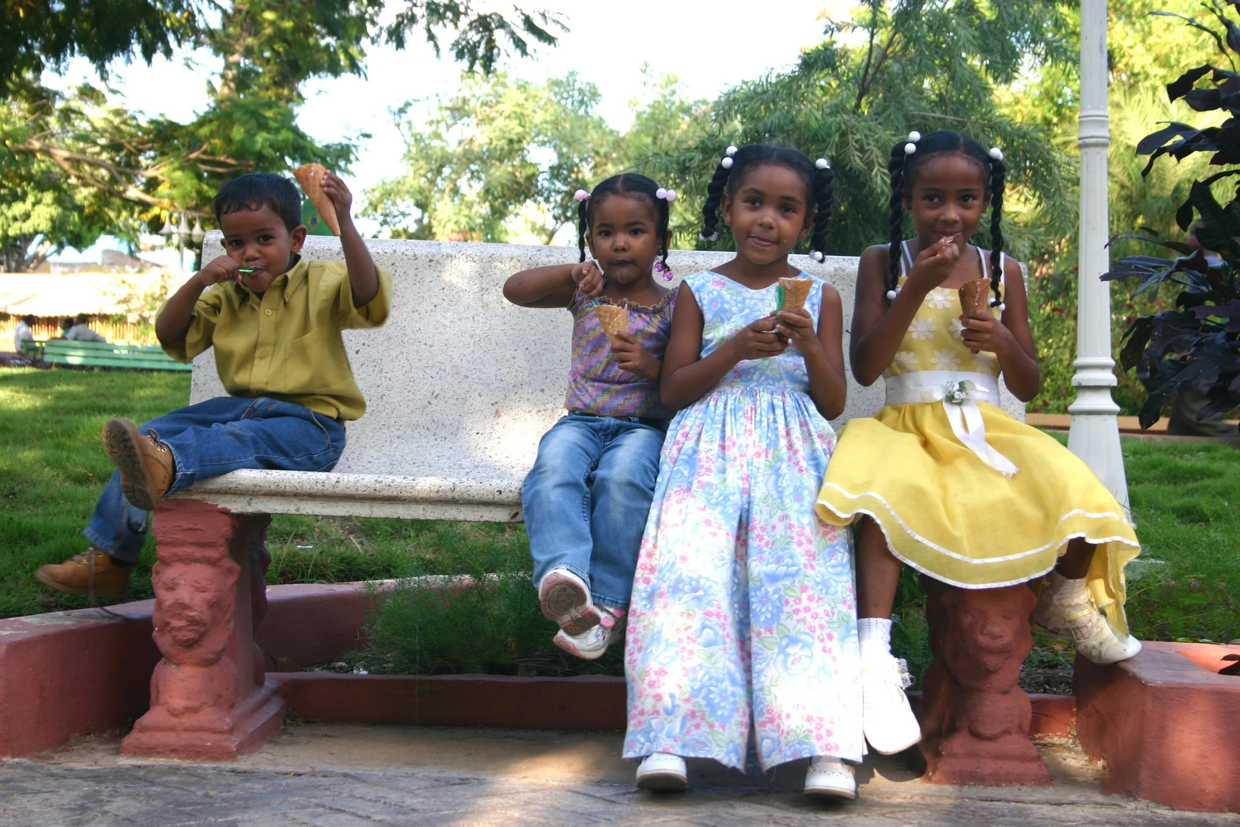 Quatre enfants assis sur un banc en pierre dans un parc, mangeant des glaces, entourés d'arbres verts.
