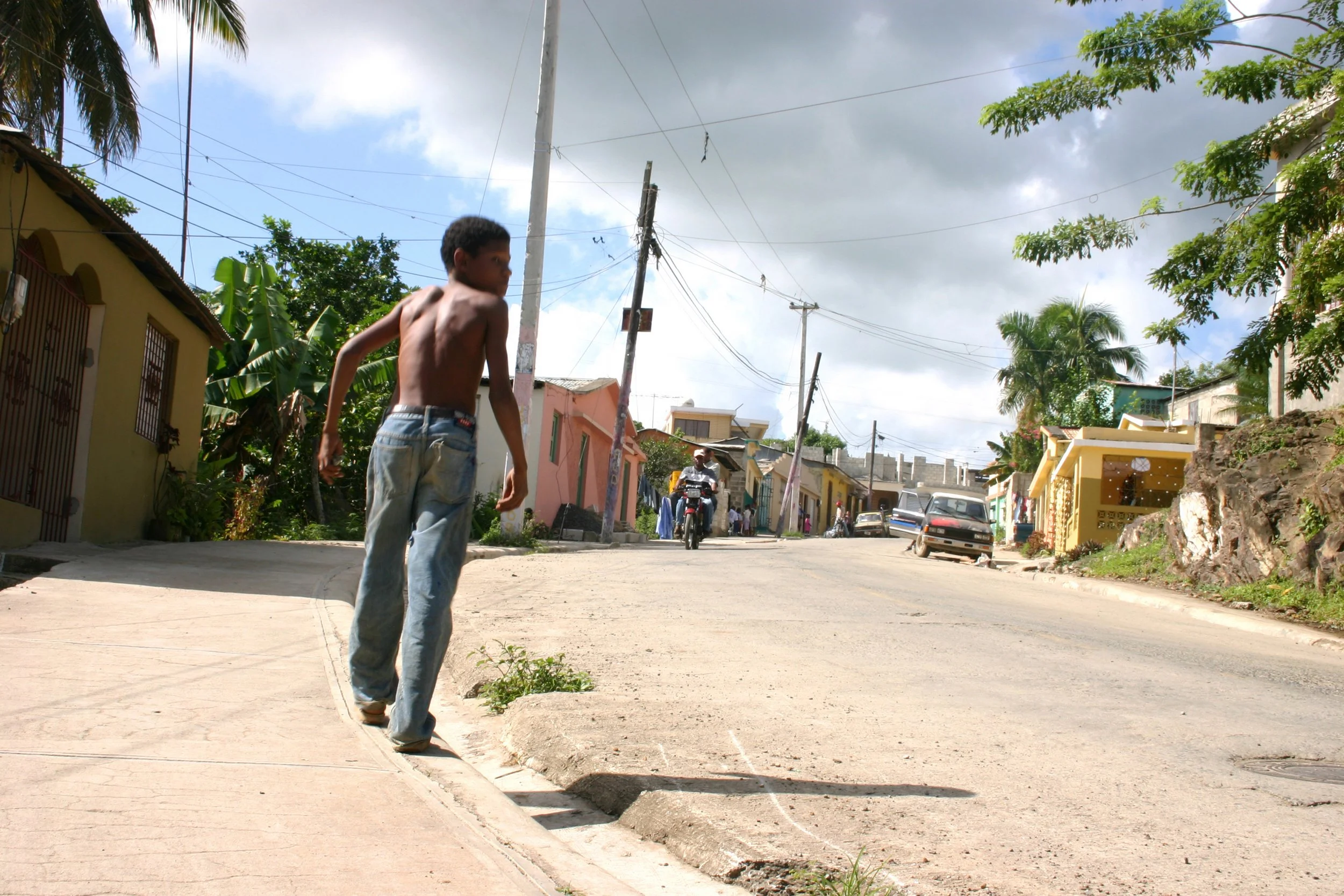Un garçon nu avec un pantalon déchiré qui marche sur le trottoir d'une rue en pente dans un quartier résidentiel tropical avec des maisons colorées, des palmiers et un ciel partiellement nuageux.