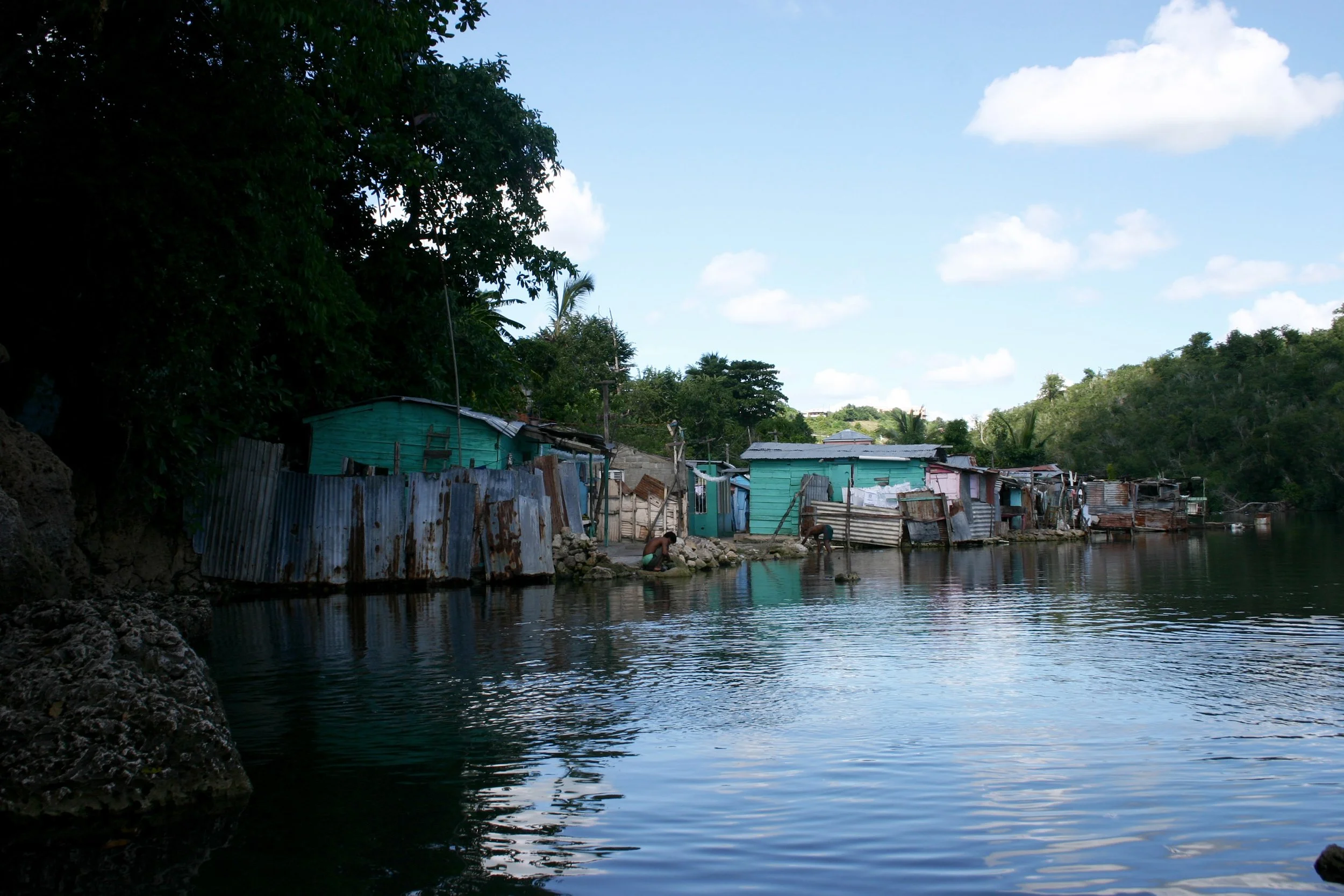 Élément de la rive d'une rivière avec des maisons en bois colorées, des gens en train de pêcher ou de laver, entourées de végétation et un ciel bleu avec quelques nuages.