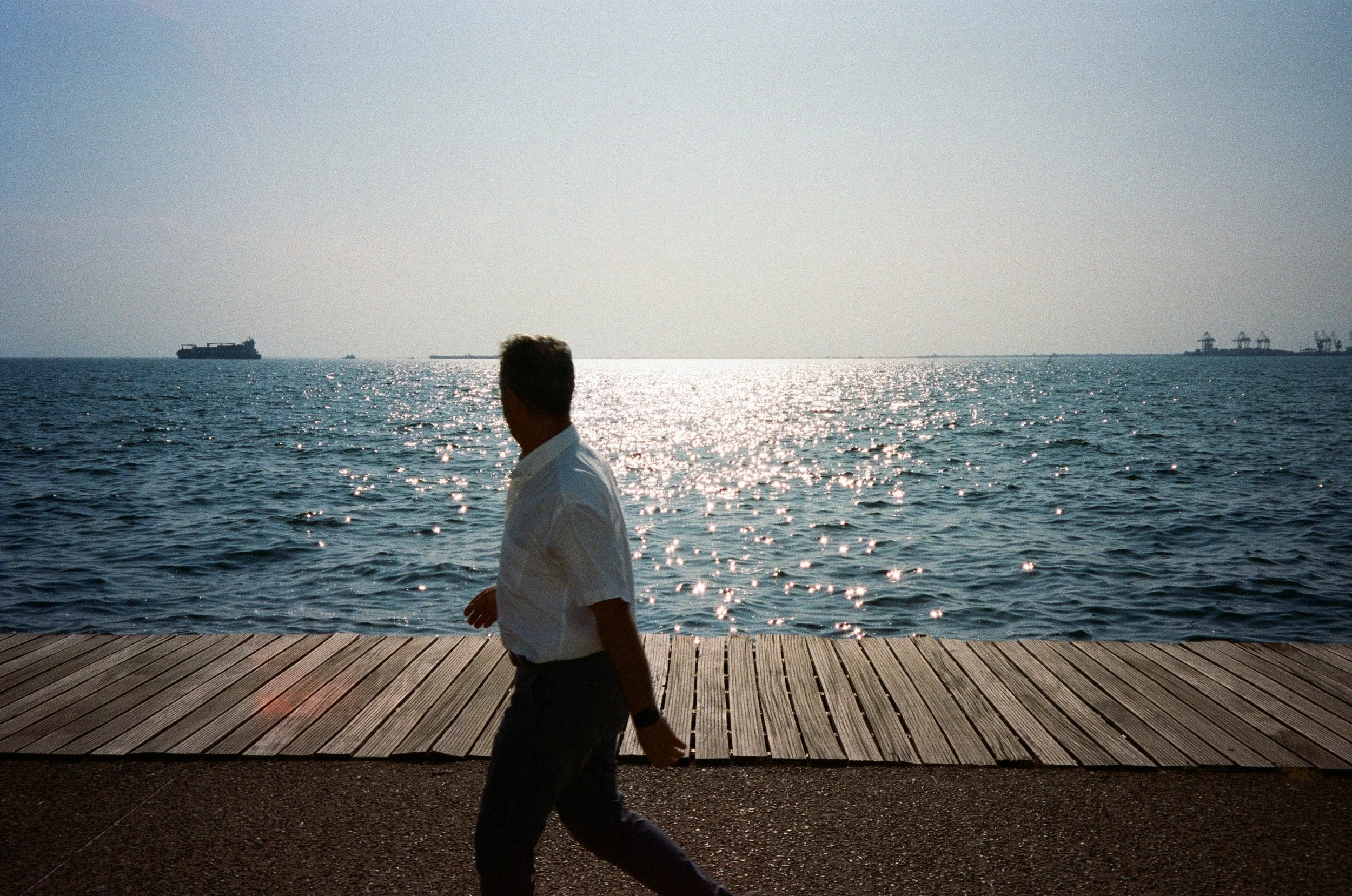Homme marchant sur une promenade en bois au bord de la mer, avec le soleil brillant sur l'eau à l'arrière-plan.