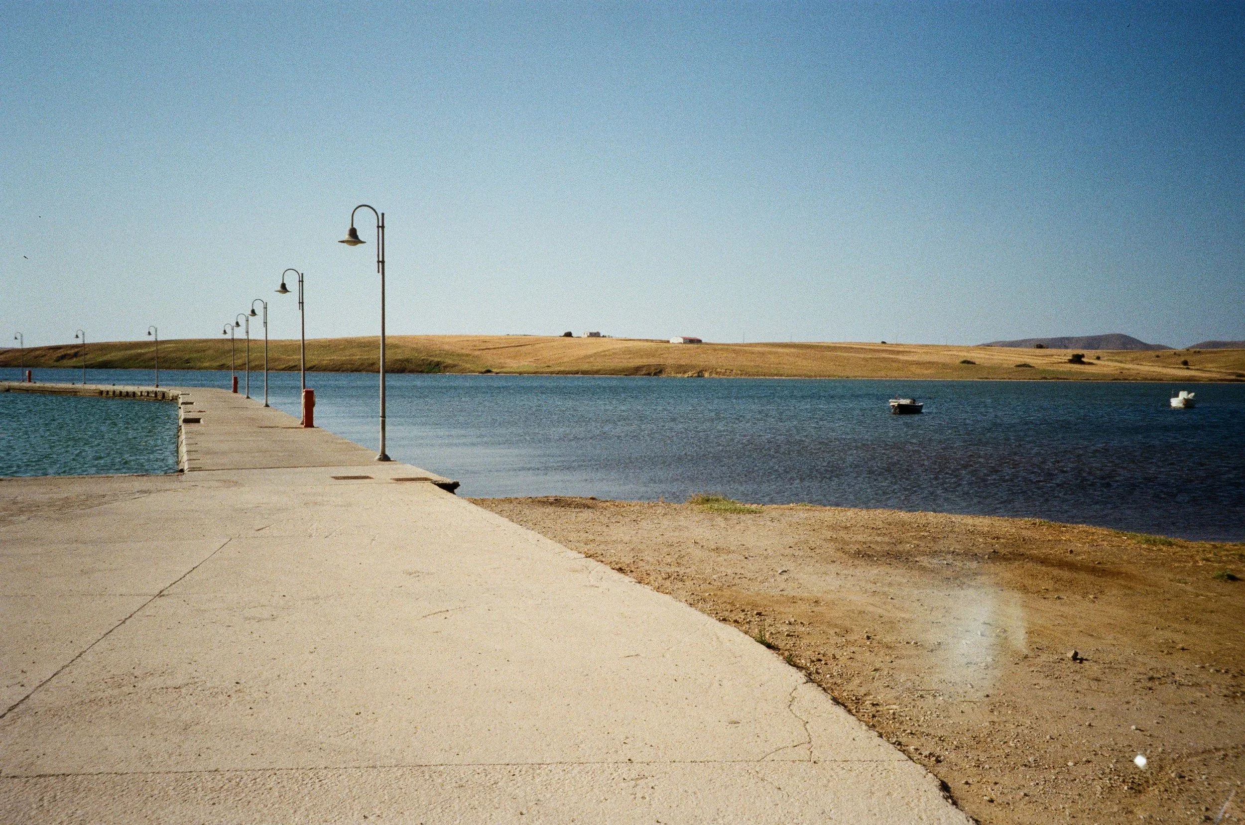 Promenade en béton longeant un lac avec des lampadaires alignés. La scène se déroule sous un ciel clair, avec une eau calme et quelques bateaux à l'ancre. Au loin, des collines ou plaines rurales s'étendent.