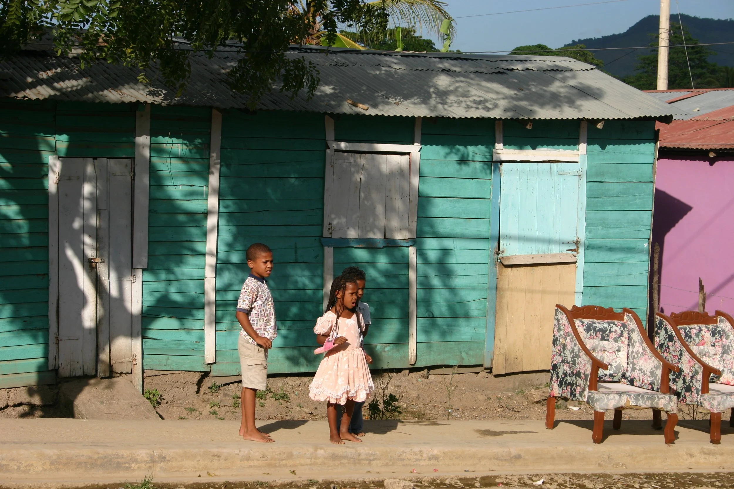 Deux enfants debout sur le trottoir devant une maison en bois peint en vert avec des fenêtres et des portes en bois blanc et coloré, sous un arbre. À droite, deux chaises avec des coussins fleuris. En arrière-plan, montagnes et lignes électriques.
