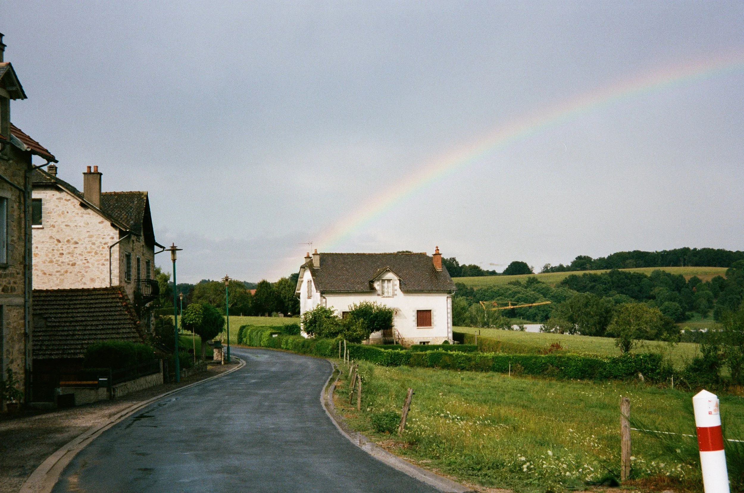 Une route de campagne bordée de maisons en pierre et de lampadaires, avec un ciel nuageux et un arc-en-ciel visible en arrière-plan au-dessus de champs verdoyants.