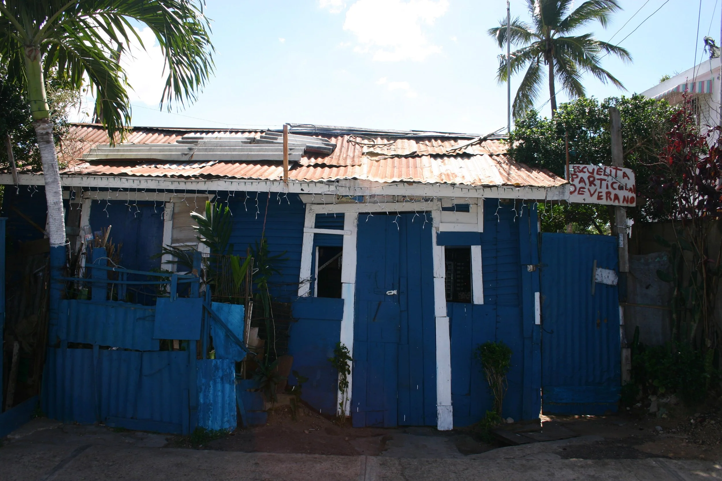 Petite maison en bois peinte en bleu avec des cadres blancs, un toit en tôle ondulée rouillée, entourée de plantes, sous un ciel clair, avec une pancarte annonçant une école d'été.