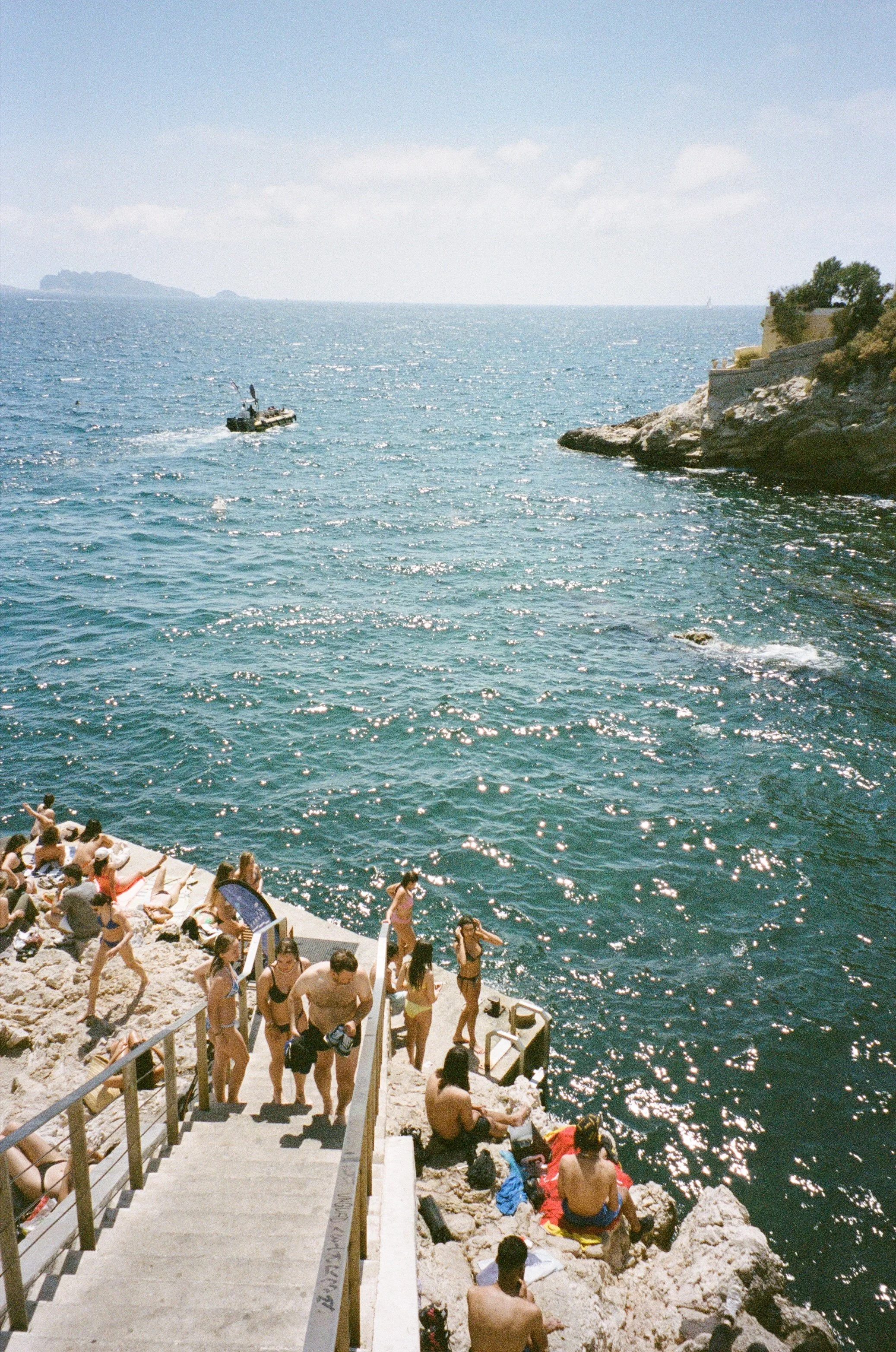 Plage en bord de mer avec des gens qui se baignent et se relaxent, bateau naviguant au loin, rochers et falaise à droite, ciel en partie nuageux.