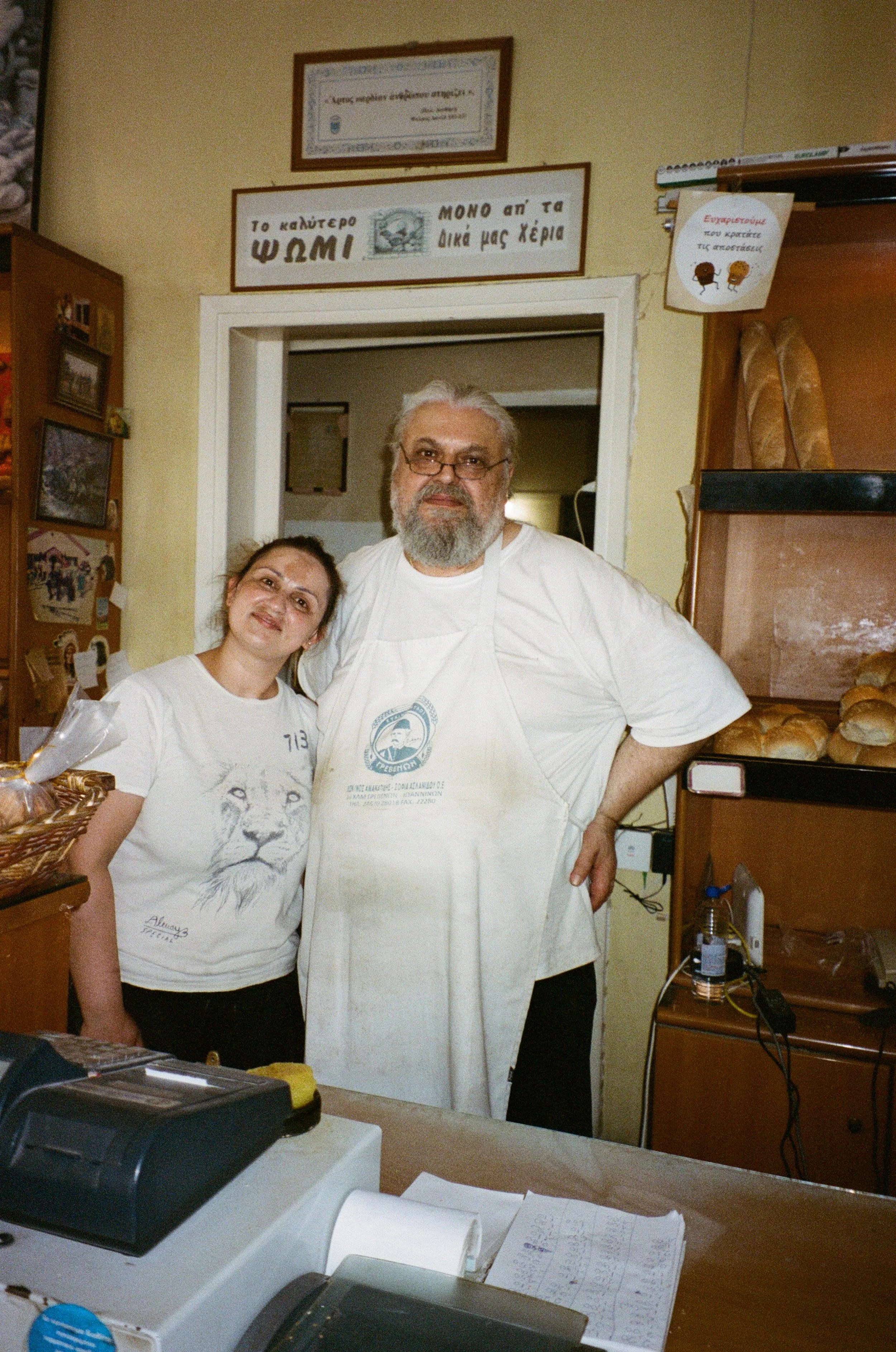 Deux personnes, un homme et une femme, posent derrière un comptoir dans une boulangerie. L'homme porte un tablier blanc avec un logo et la femme un t-shirt blanc avec un lion. Il y a des pains sur une étagère à droite et des affiches en grec au mur.