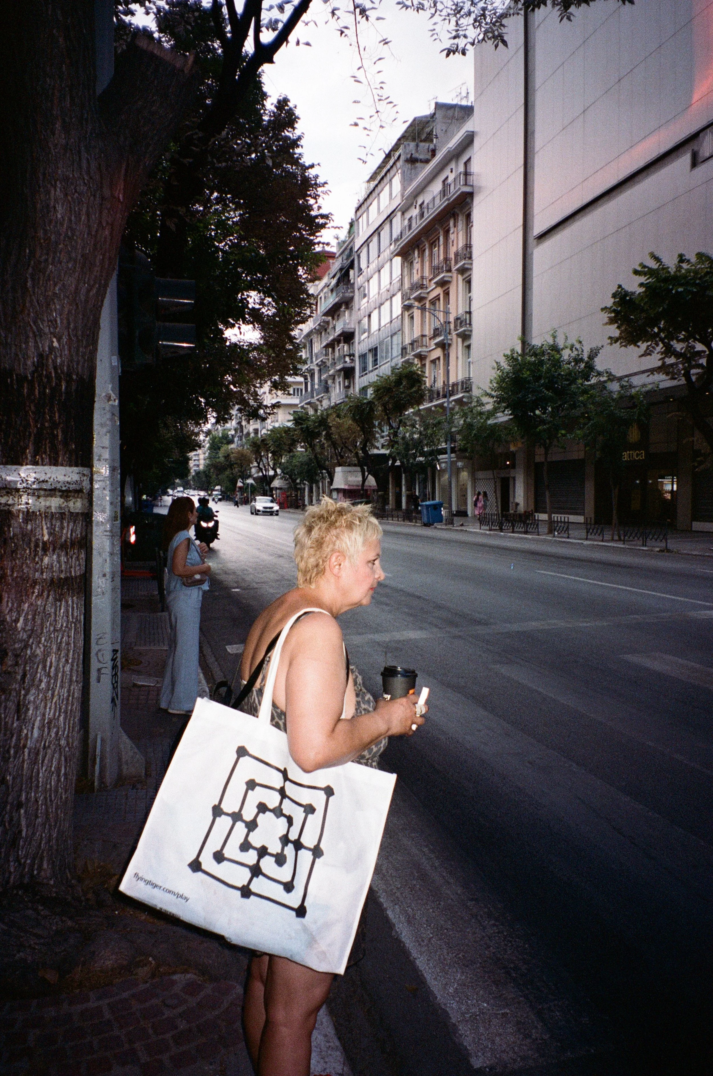 Une femme aux cheveux blonds courts portant un porte-sac blanc avec un motif noir, tenant une tasse de café et une cigarette, attendant sur le trottoir d'une rue urbaine.