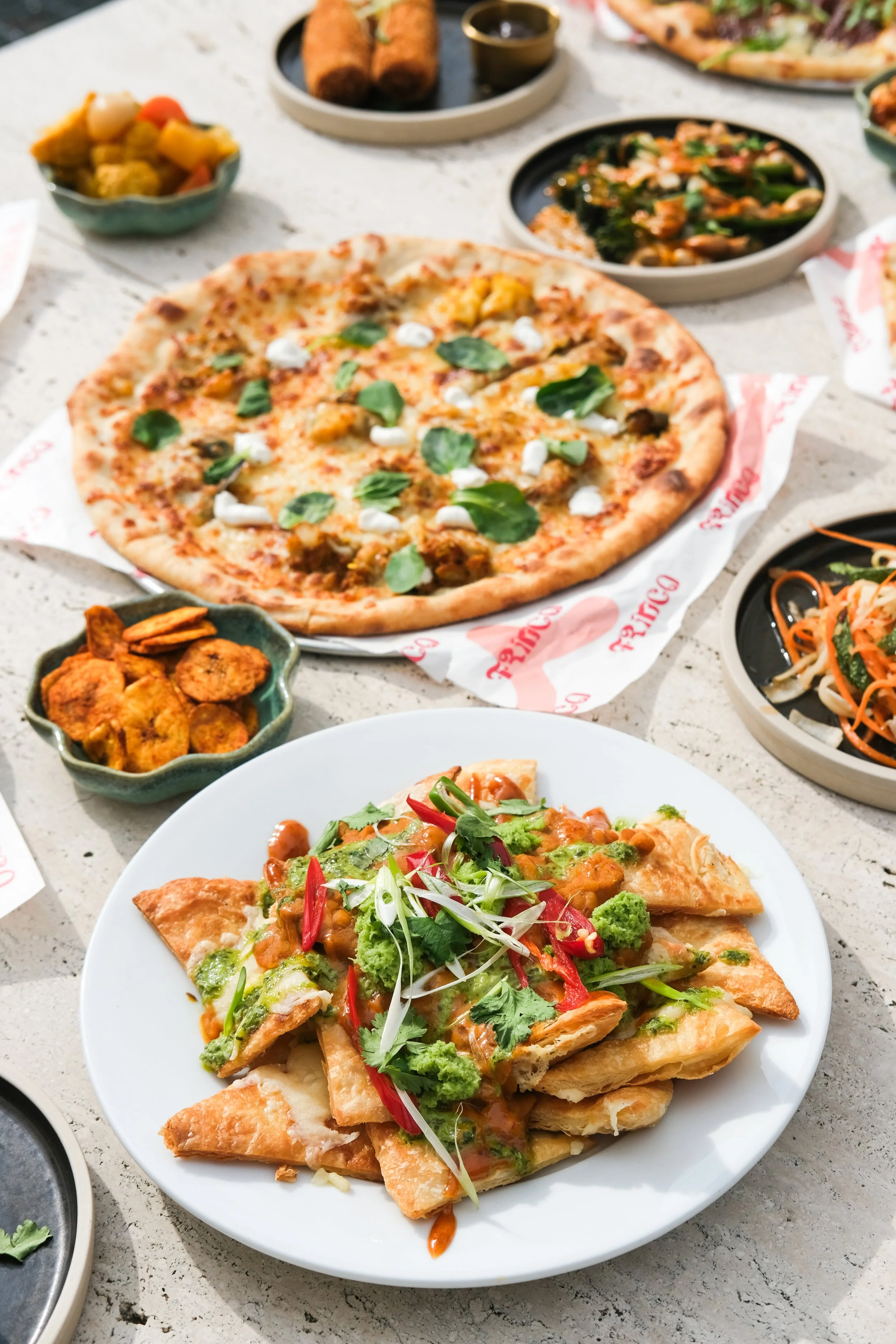 Pizza with green leaves and cheese, fried dumplings, salad with vegetables, and other Asian dishes on a table.