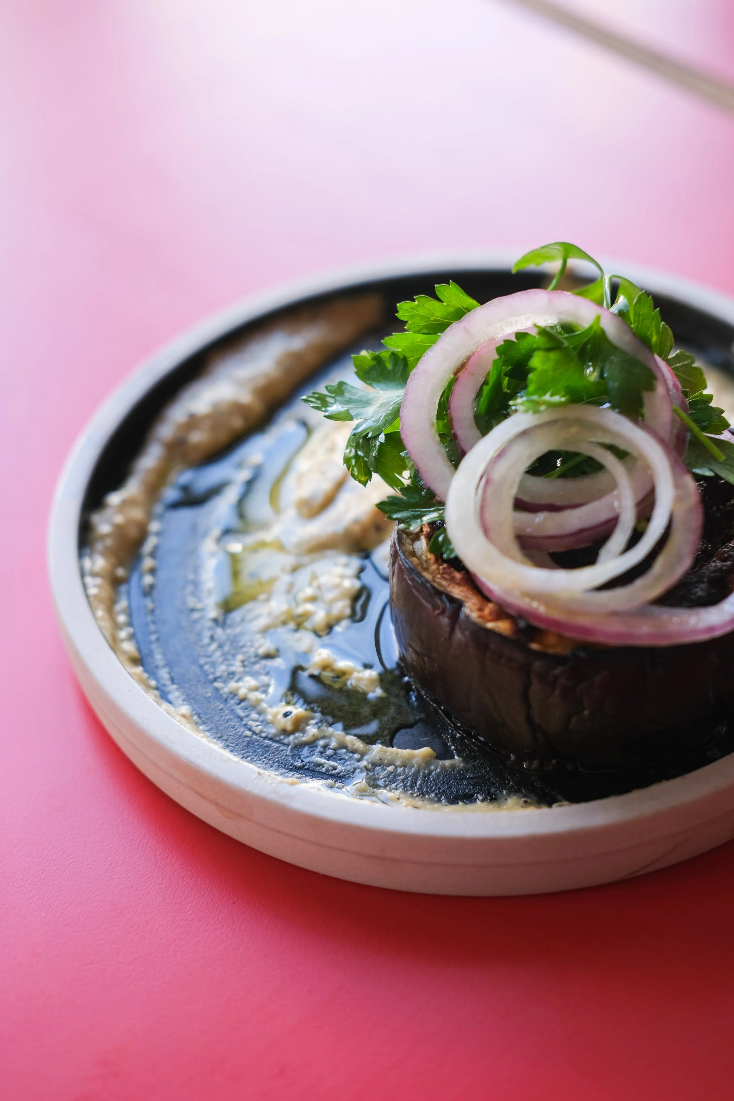 Baked eggplant topped with sliced onions and parsley, served with a sauce or dip in a bowl, on a pink surface.