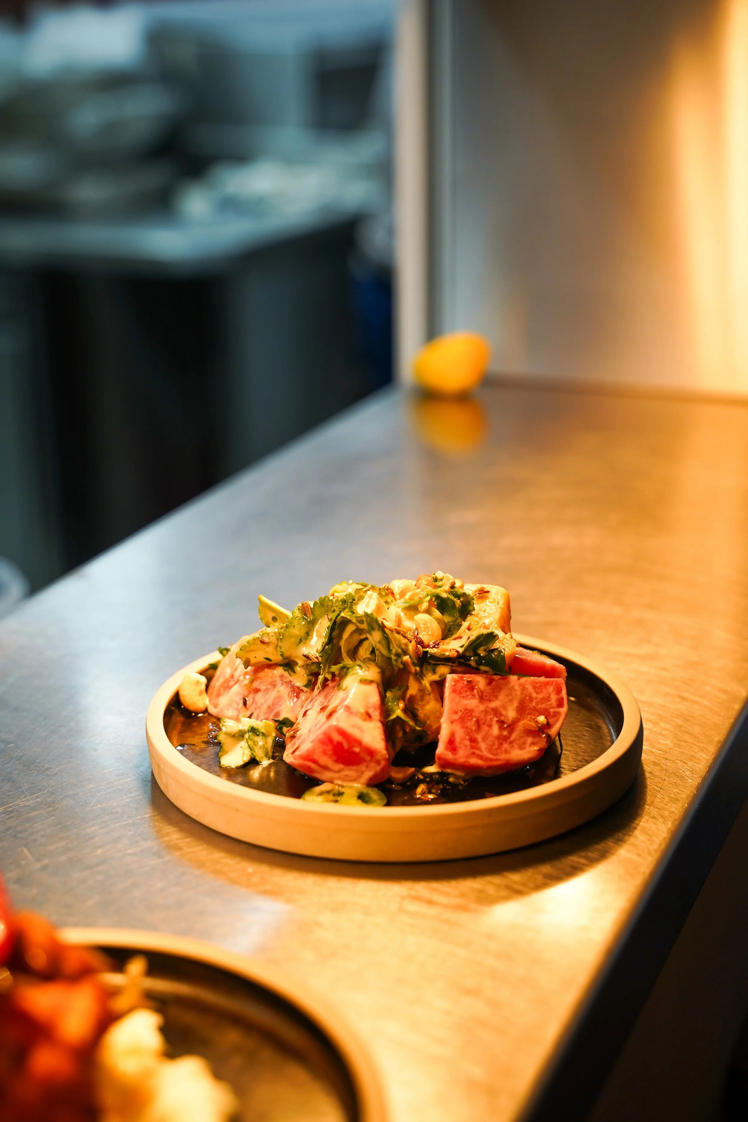 Raw tuna sashimi served on a black plate with seaweed and sesame seeds, placed on a wooden dish on a stainless steel countertop.