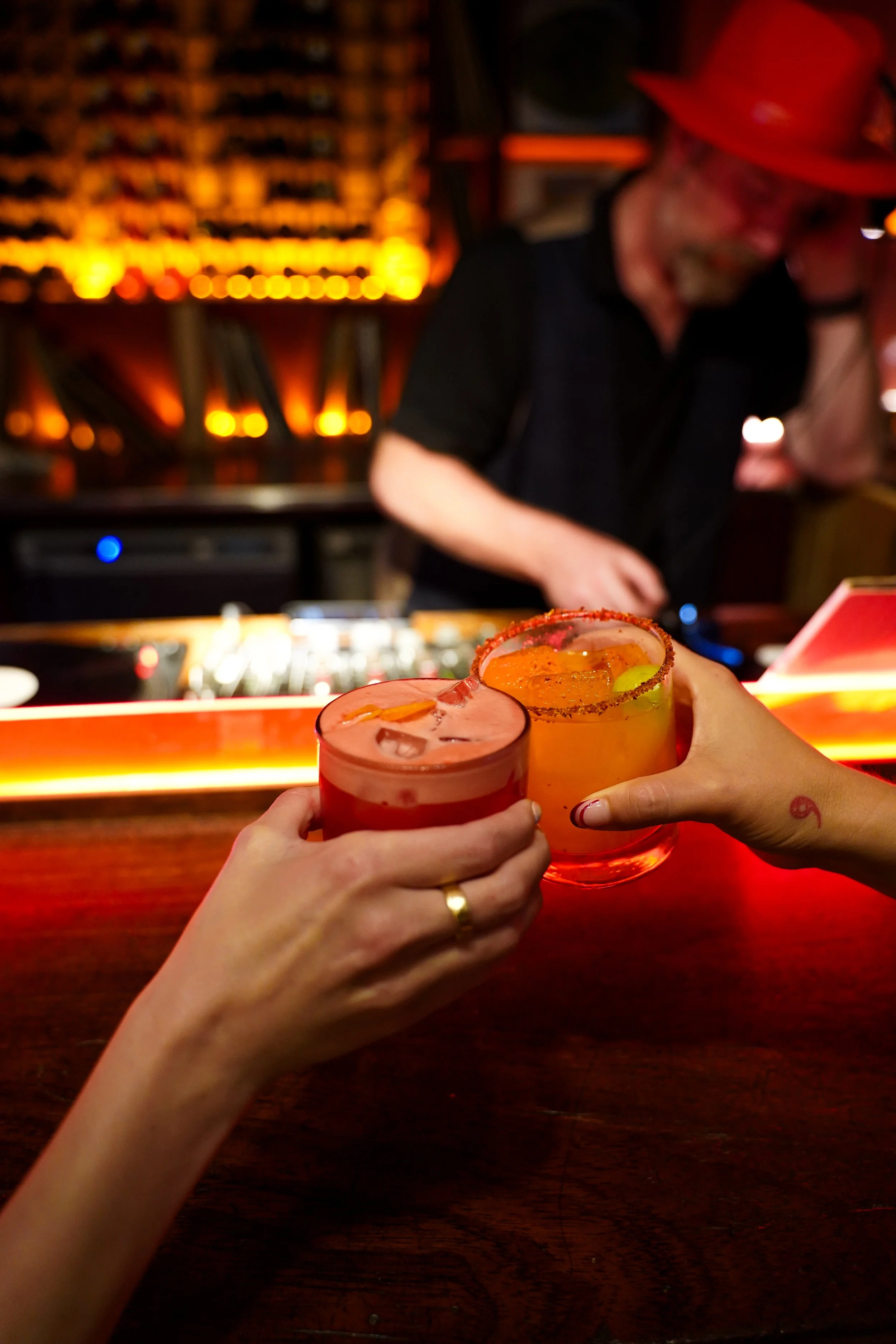 Two hands holding colorful cocktails with salted rims, raised in a toast at a bar, with a DJ in the background.