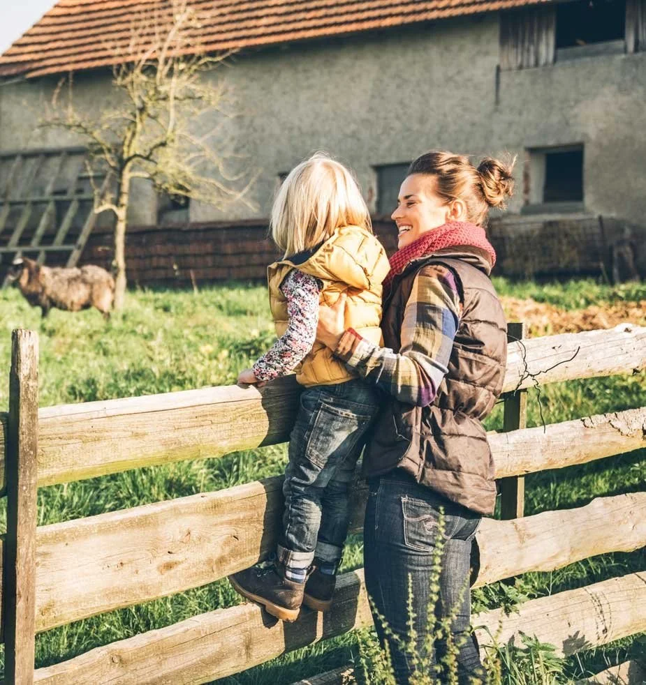 A woman and a young girl smiling and enjoying time together at a farm, with a goat in the background near a wooden fence.