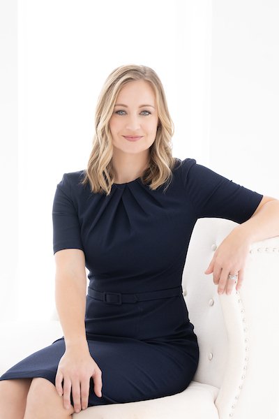 Clair Jody, Administration and Finance Leader at Warehouses4Good, with blonde hair, wearing a navy blue dress, sitting on a white chair with her right arm resting on the armrest, smiling at the camera.