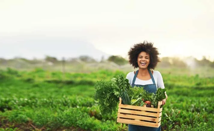 A woman with curly hair smiling while holding a wooden crate filled with fresh vegetables outdoors in a farm field.