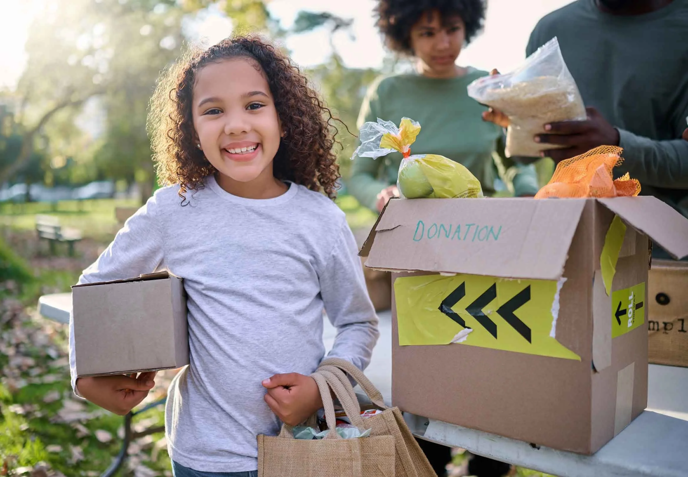 A young girl smiling and holding a donation box outdoors. Behind her are two people sorting donations into a cardboard box labeled "Donation" on a table. The scene takes place in a park with trees and sunlight.