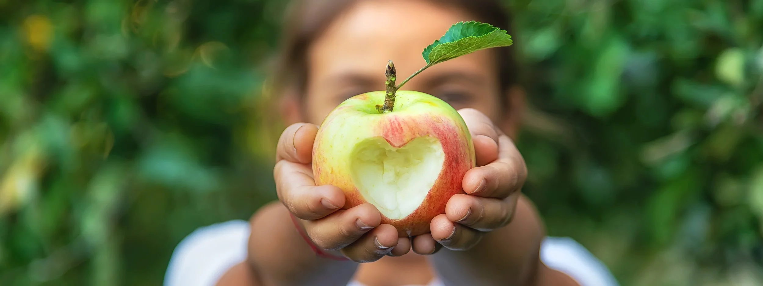 Girl holding a partially eaten apple with a leaf attached, outdoors with green foliage in the background.