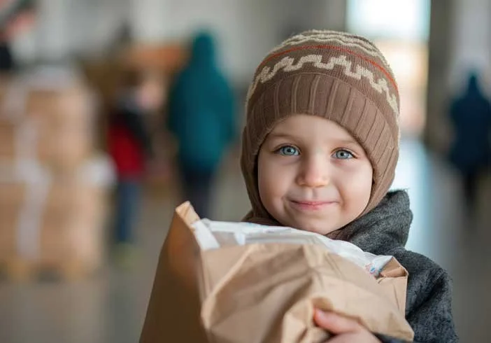 A young boy with blue eyes wearing a brown knit hat and a dark gray coat, holding a paper bag in a busy indoor setting.