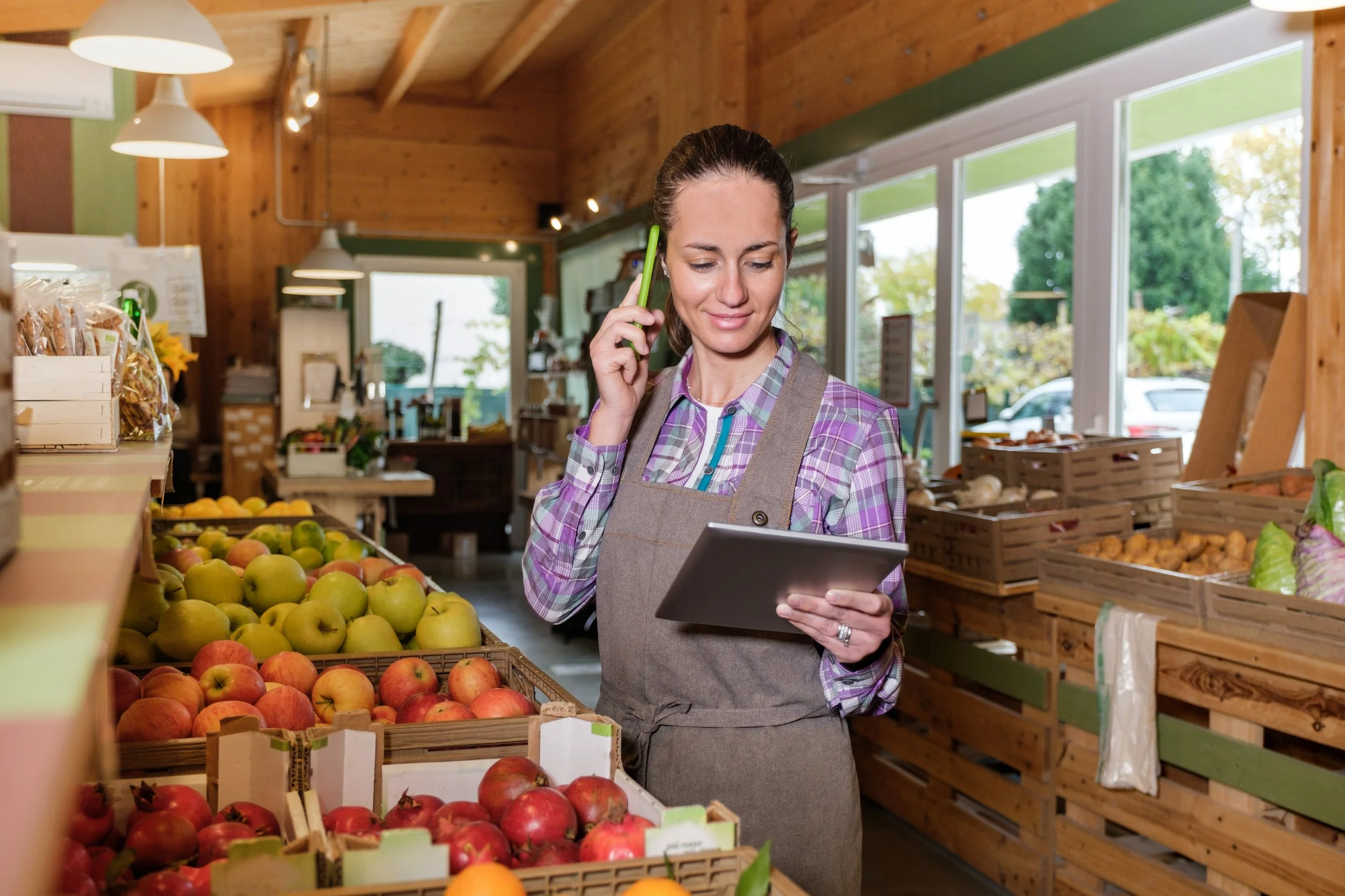 A woman in a plaid shirt and apron inside a grocery store, talking on her phone while looking at a tablet, surrounded by crates of fruits and vegetables.
