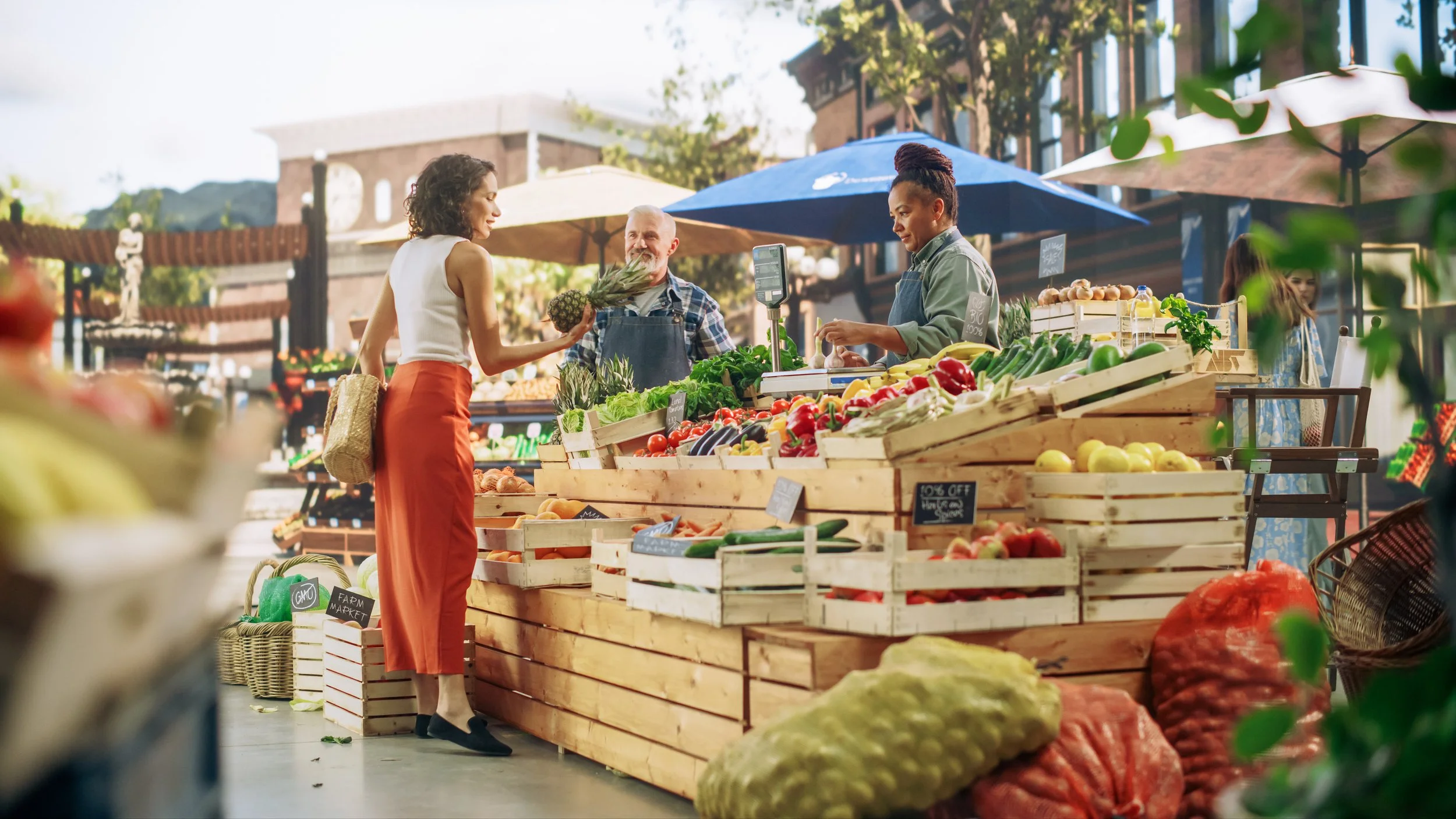 An outdoor farmers market with vendors selling fresh fruits and vegetables, and a woman shopping with two vendors under umbrellas.
