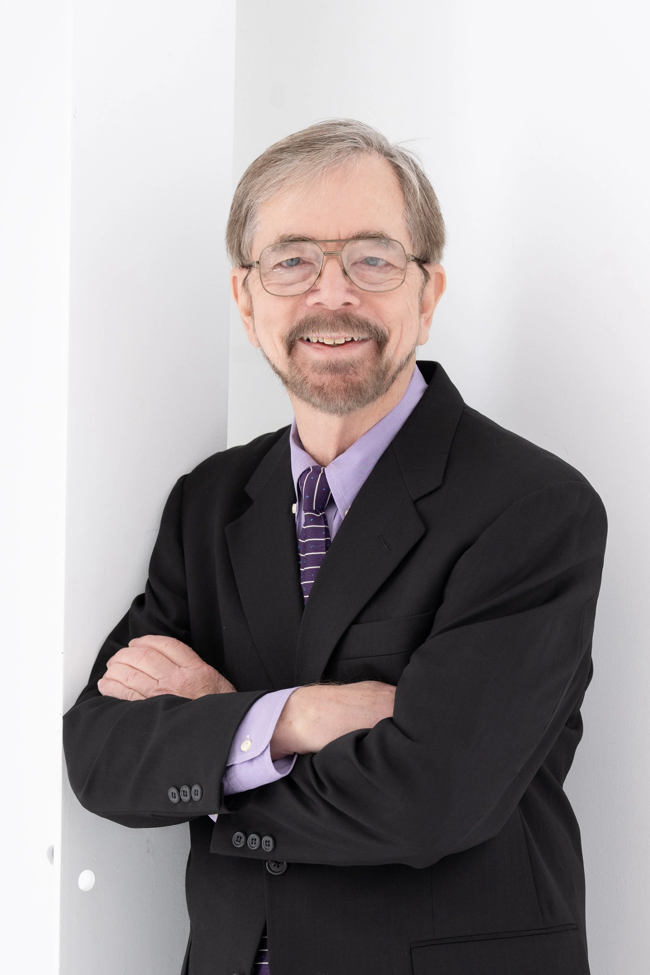 John Kane, Executive Director at Warehouses4Good, wearing a black suit, purple dress shirt, and striped tie, standing with arms crossed against a white wall.