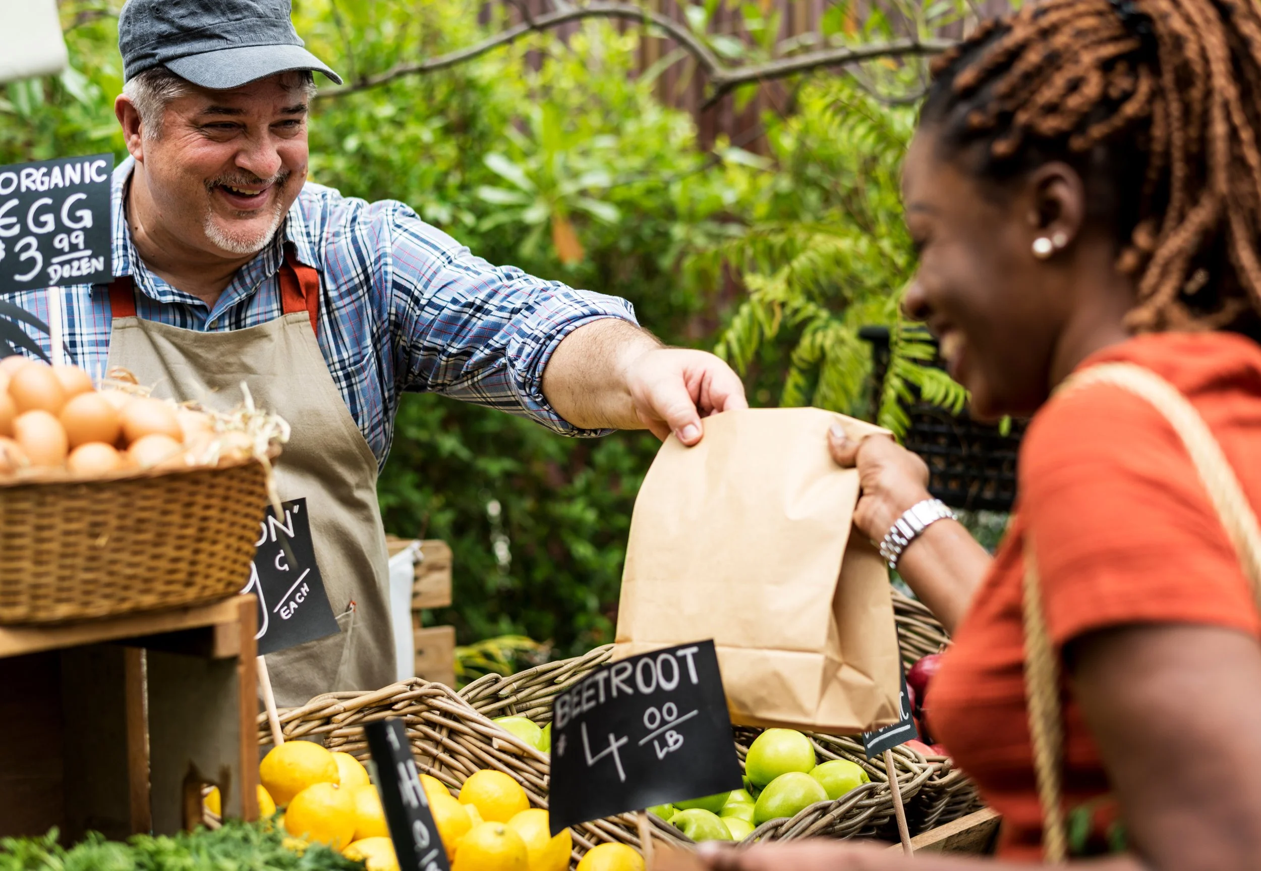 A smiling man selling organic eggs at a farmers market, handing a paper bag to a woman customer with fresh vegetables and produce around them.