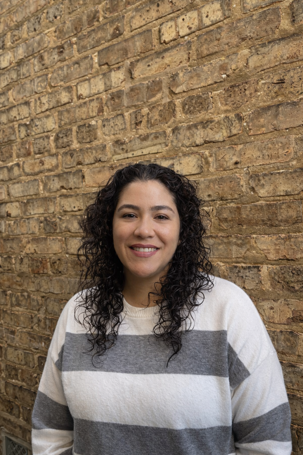 A woman with curly dark hair, smiling, wearing a cream and gray striped sweater, standing in front of a rustic brick wall.