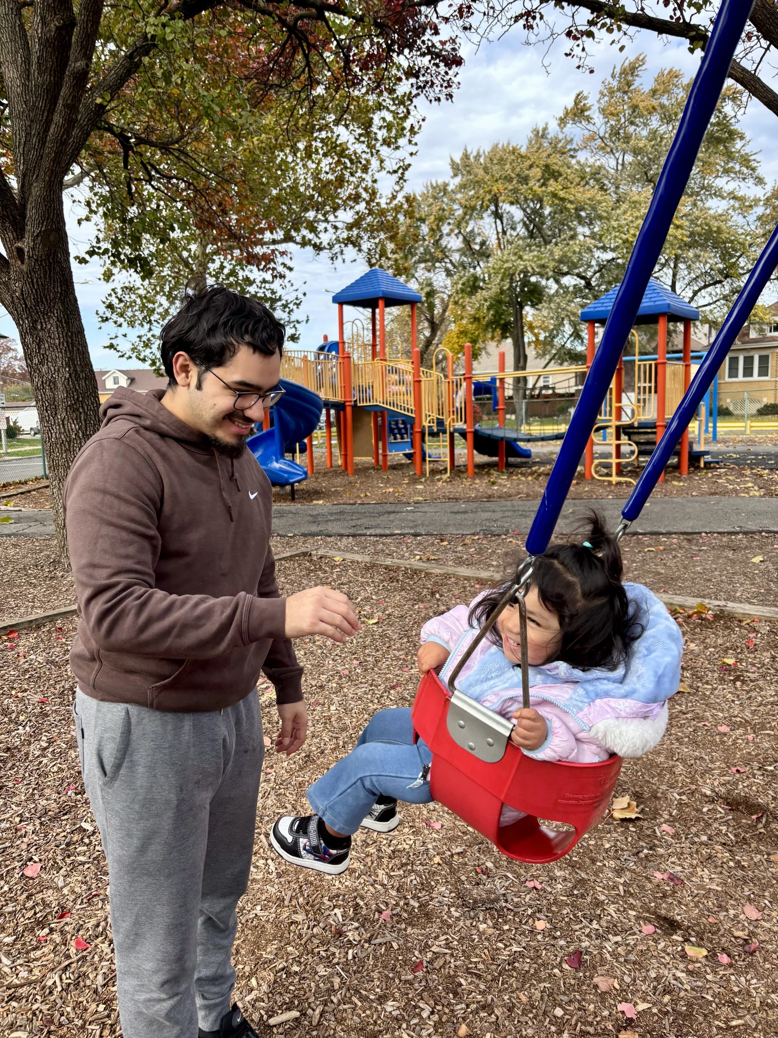 A man with glasses playing with a young girl sitting in a red swing at a playground, with trees and playground equipment visible in the background on a fall day.