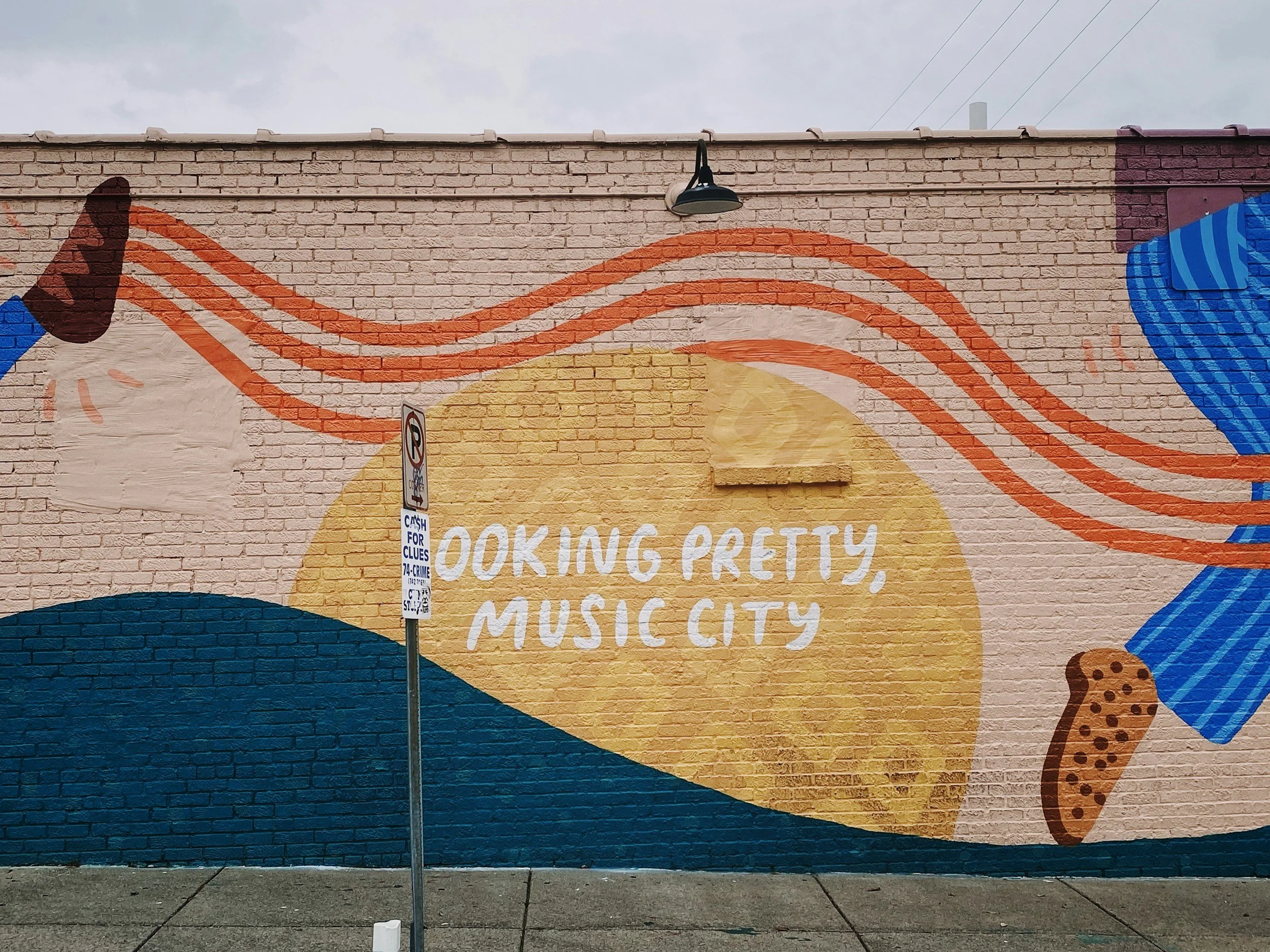 Colorful mural on a brick wall featuring an orange sun, blue waves, and stylized ice cream cones. White text in the center reads, 'Looking pretty, Music City.'