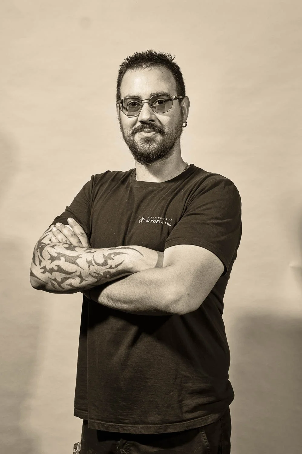 Un homme à la peau claire, portant des lunettes, un tatouage sur le bras droit, un t-shirt noir, se tient avec les bras croisés devant un mur beige.