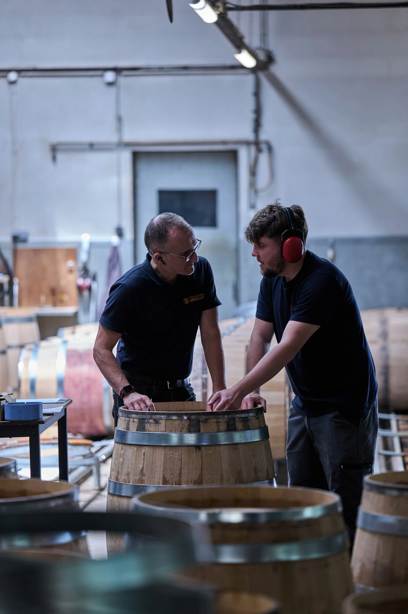 Deux hommes travaillent dans un atelier de tonnellerie, entourés de barriques en bois.