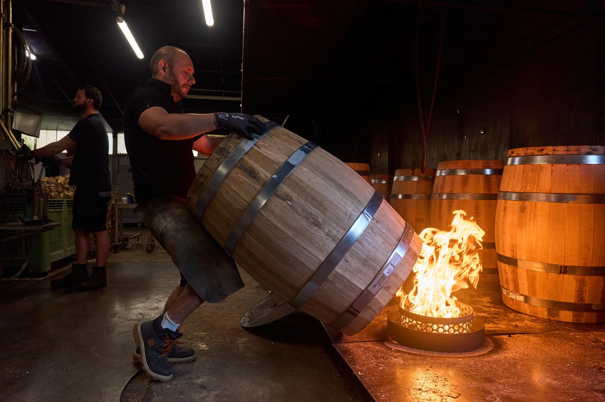 Personne en train de d'installer un tonneau en bois sur un feu dans une tonnellerie, avec d'autres tonneaux en arrière-plan.
