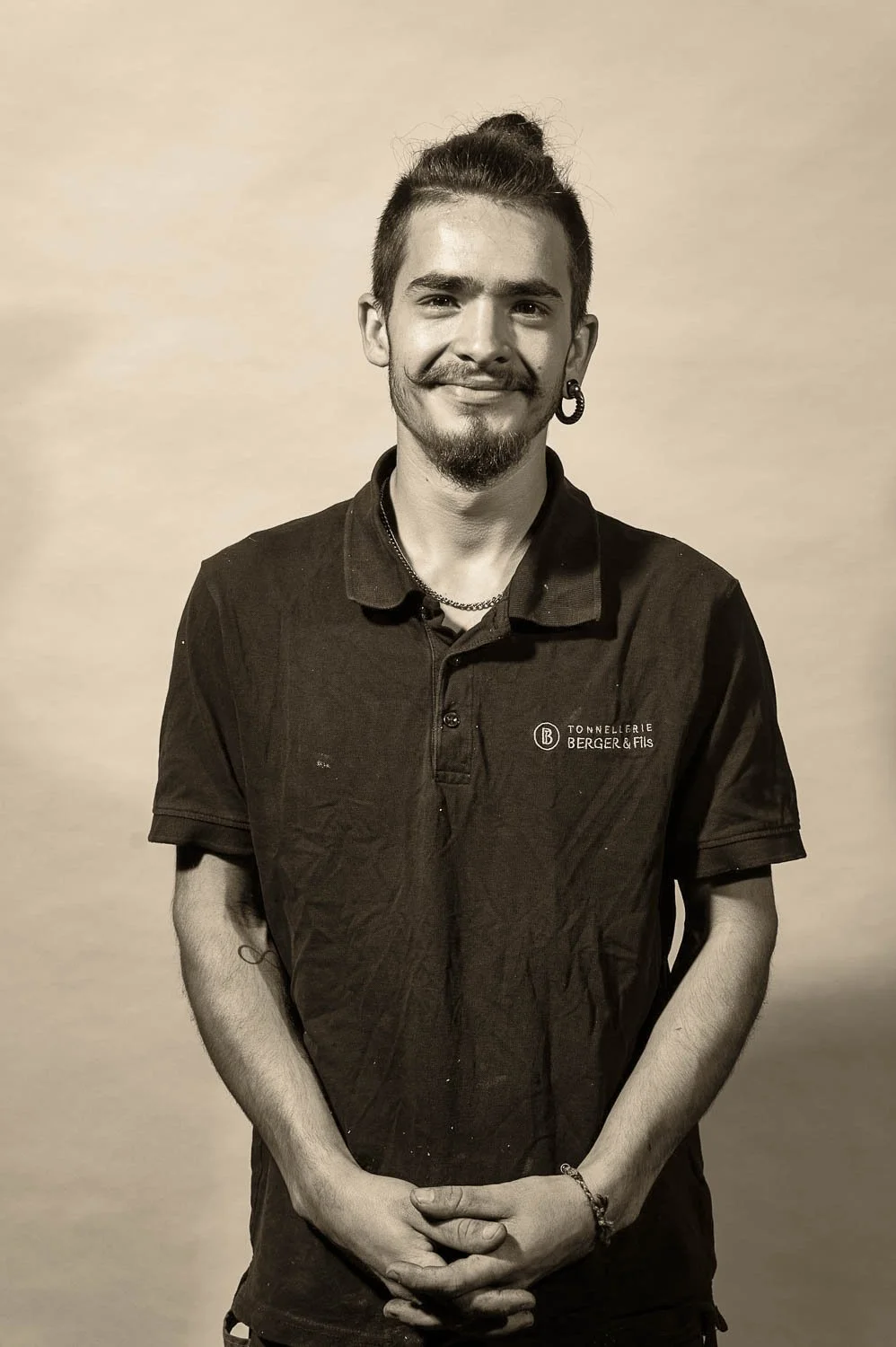 Portrait en noir et blanc d'un homme souriant avec une boucle d'oreille, portant un polo avec logo 'Tonnellerie Berger & Fils'.
