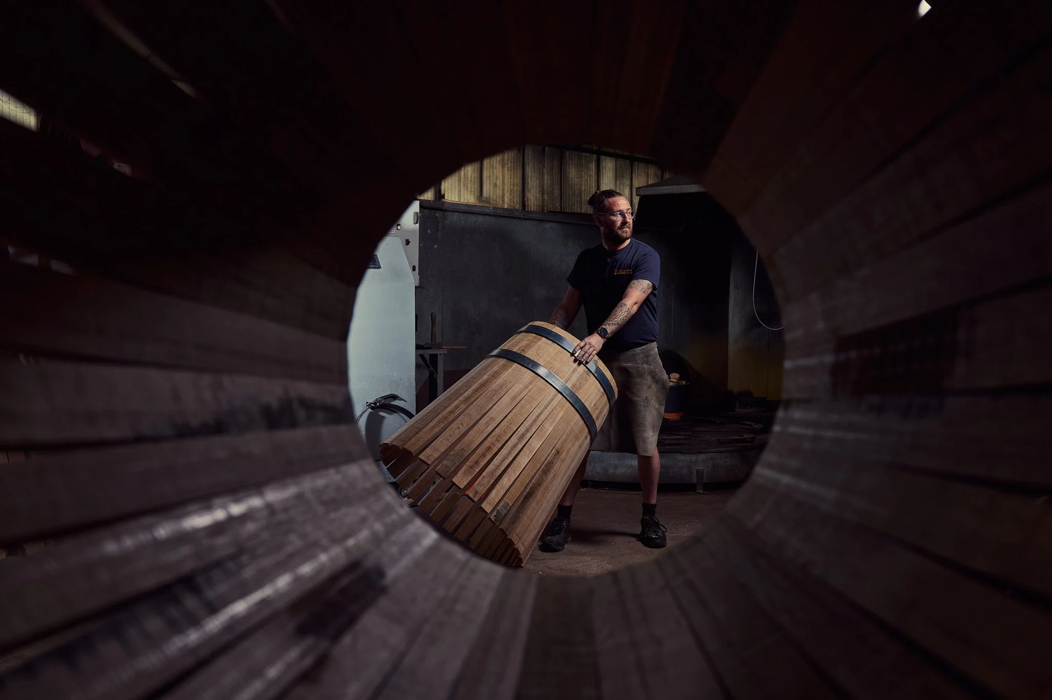 Un homme travaille dans un atelier de fabrication de tonneaux en bois, vu à travers un trou circulaire dans un tonneau.
