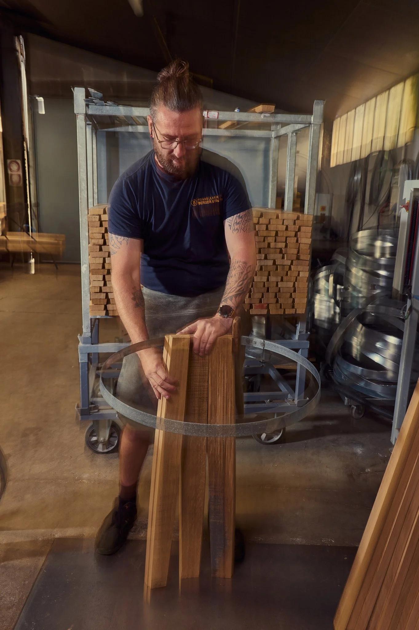 Un Tonnelier tatoué en train de couper du bois dans un atelier, avec une cheminée de bois en arrière-plan.