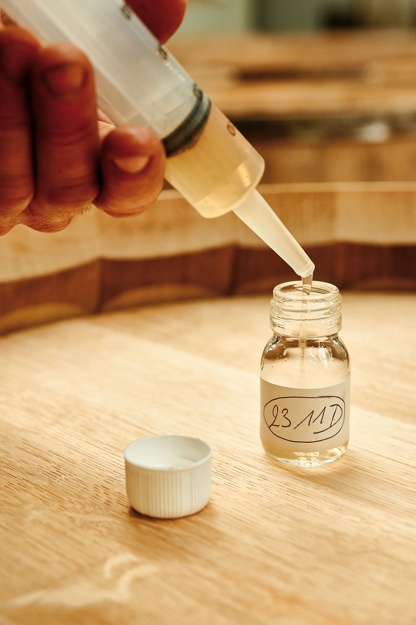 Une main verse un liquide transparent d'une seringue dans un petit flacon en verre avec une étiquette portant la date 23/11.