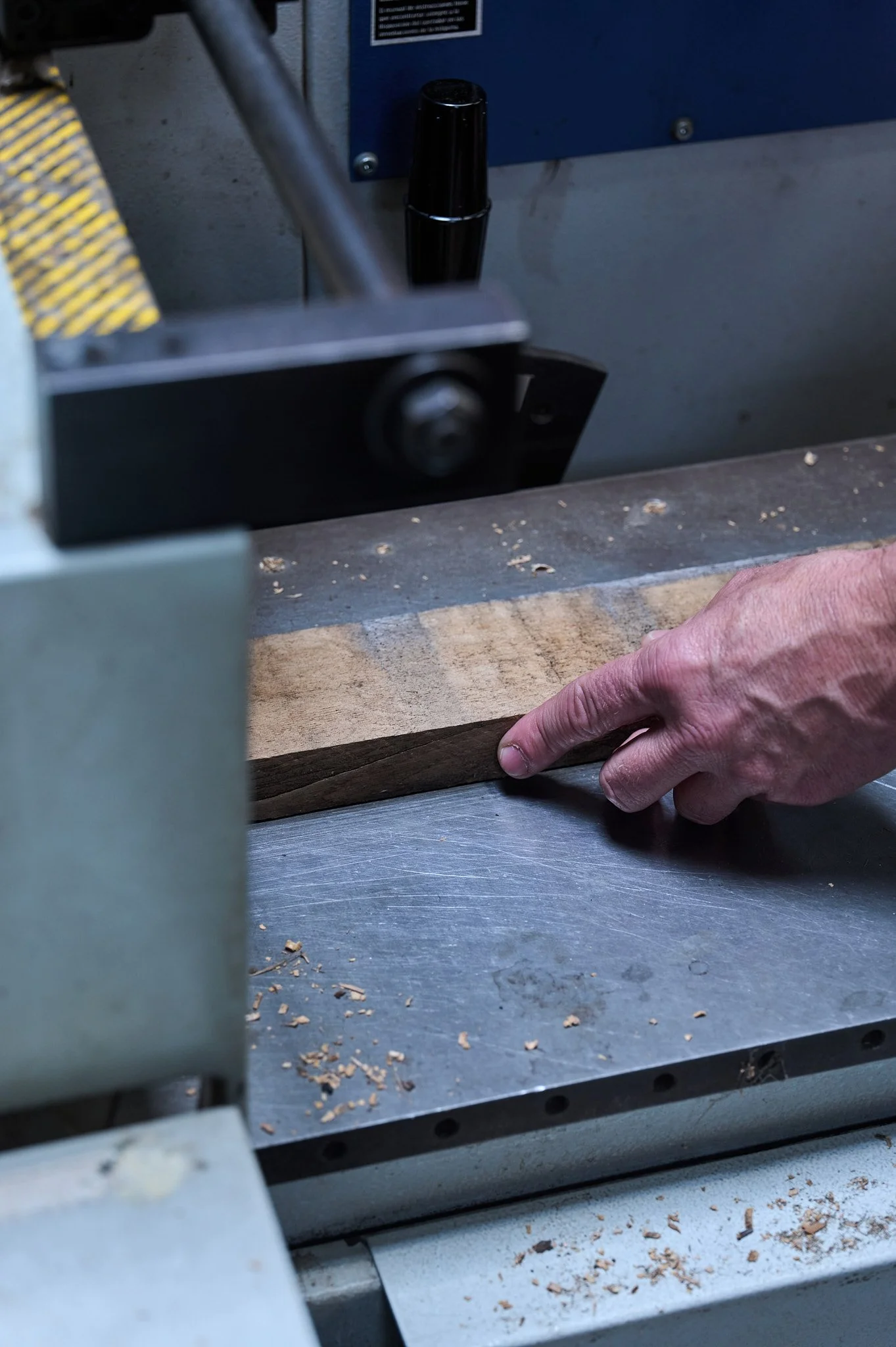 Une main manipule une pièce de bois sur une machine-outil, probablement une raboteuse ou une plane. La machine est grise avec quelques copeaux de bois autour.